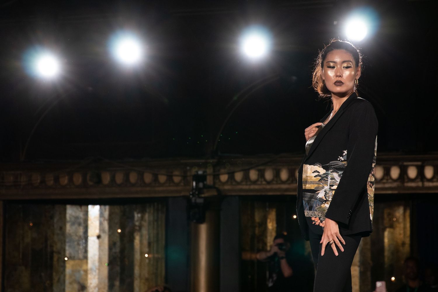 Model poses in black outfit with decorative panel under stage lights at fashion show.