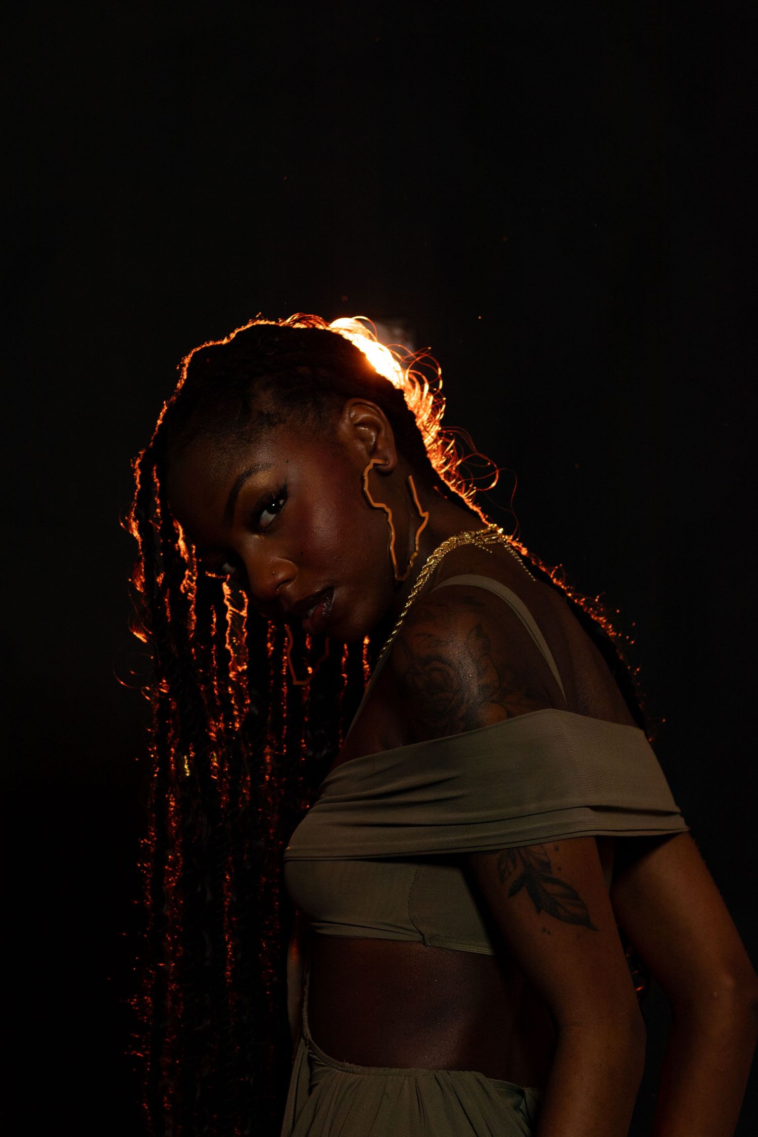 Black woman with long braids, shoulder tattoo, and gold jewelry posing in dramatic backlit warm lighting.