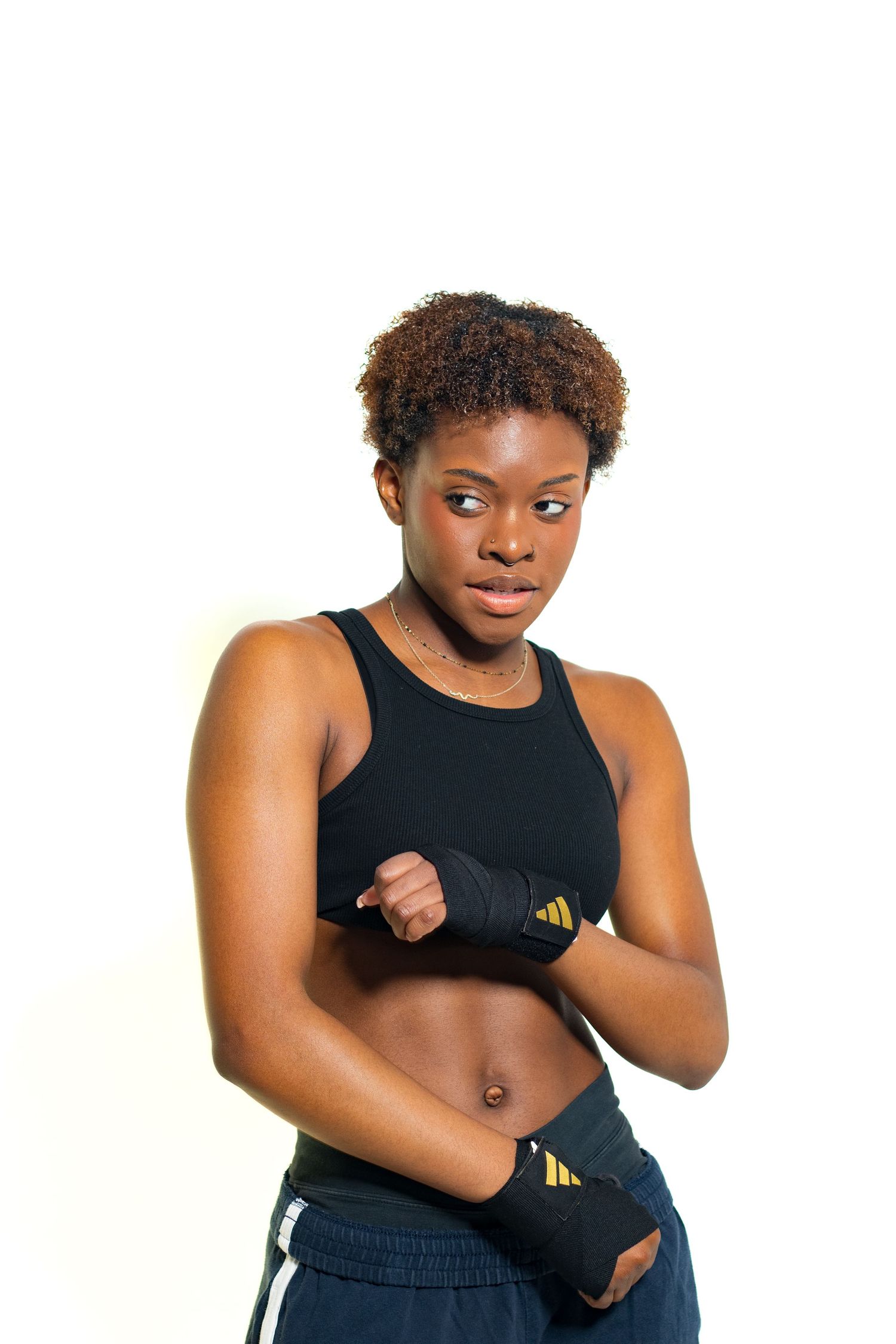 Fit Black woman in boxing hand wraps and sports bra in defensive fighting stance on white background.