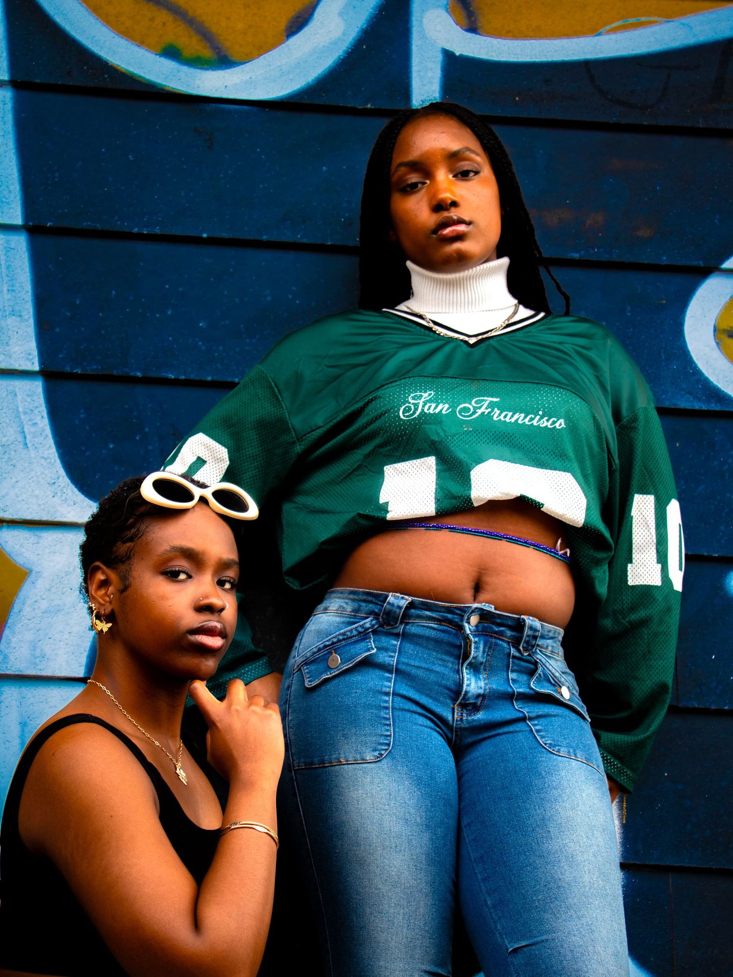 Two stylish young women posing against a blue graffiti wall in urban outfits.
