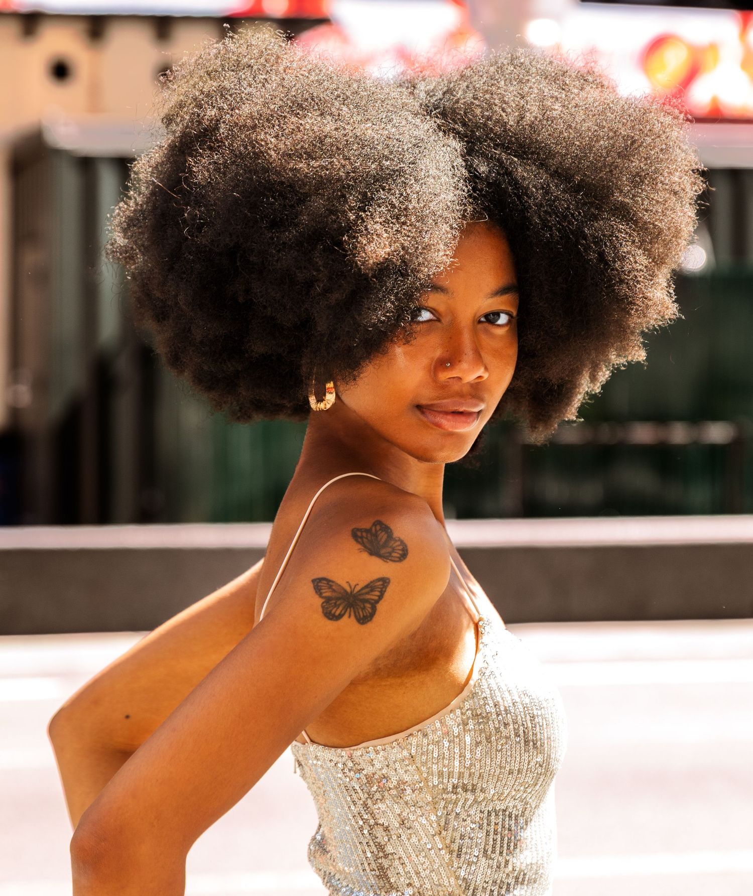 Young Black woman with large natural afro hairstyle wearing silver sequin dress with butterfly tattoo on shoulder.