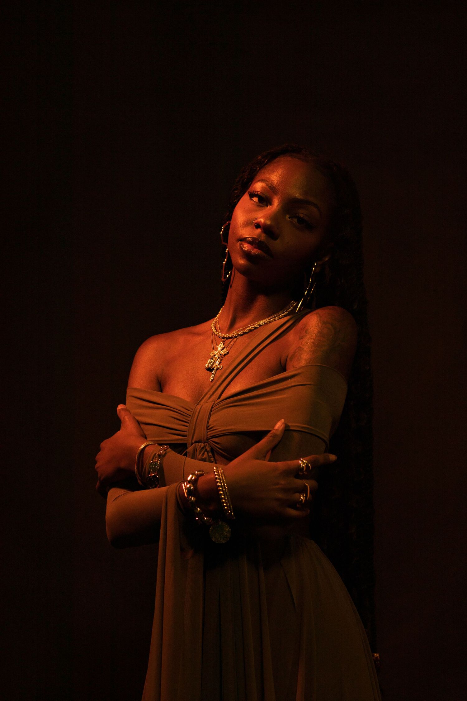 Black woman in olive off-shoulder dress with gold jewelry, arms crossed, posing against dark background.