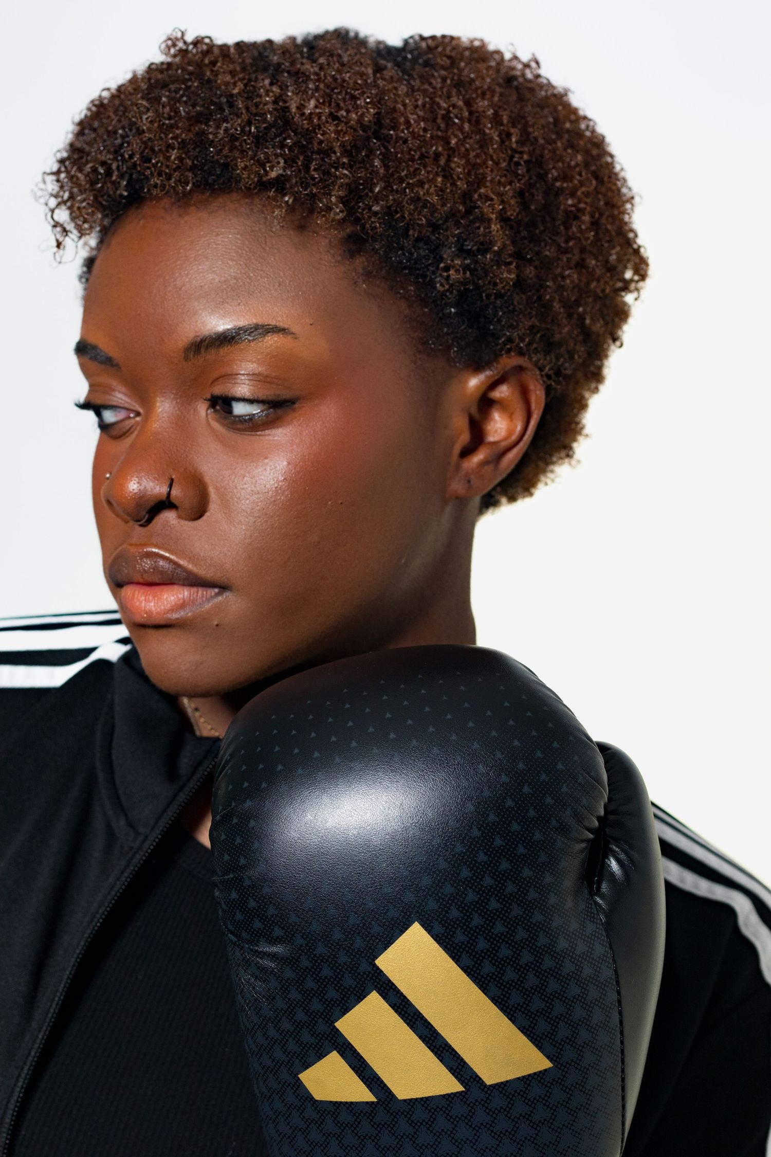 Close-up side portrait of a young Black female boxer in Adidas jacket with gold-logo boxing glove.