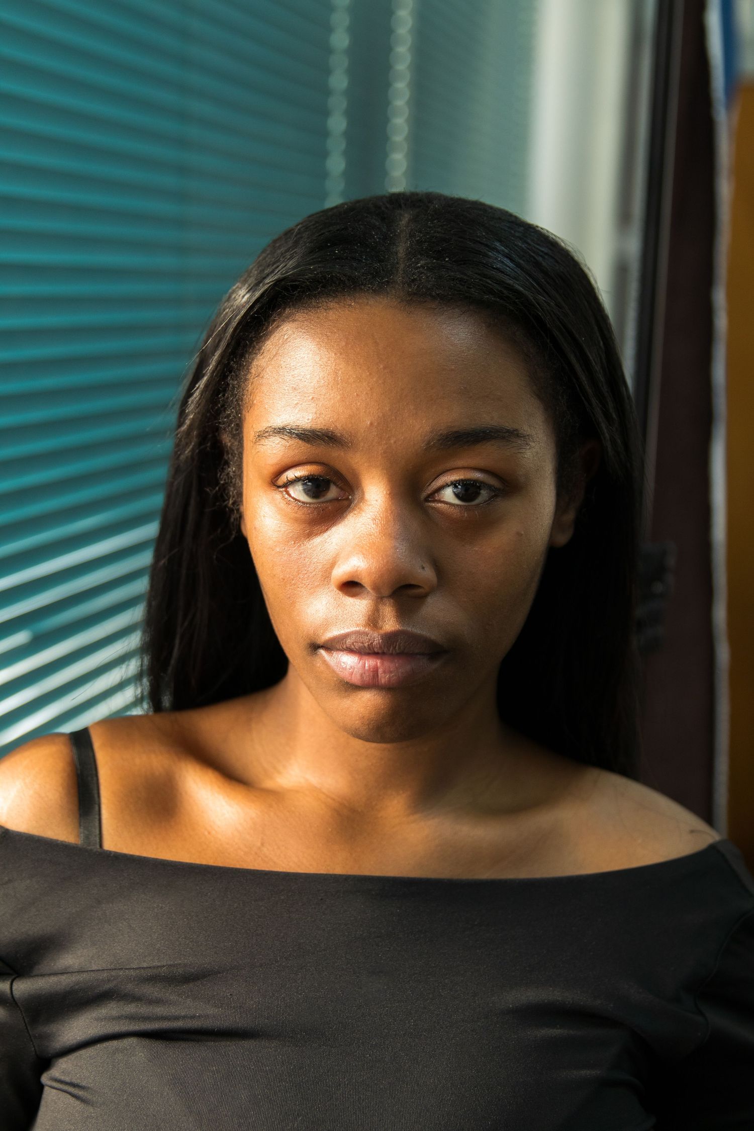 Close-up portrait of a Black woman with straight hair wearing a black off-shoulder top near window blinds.