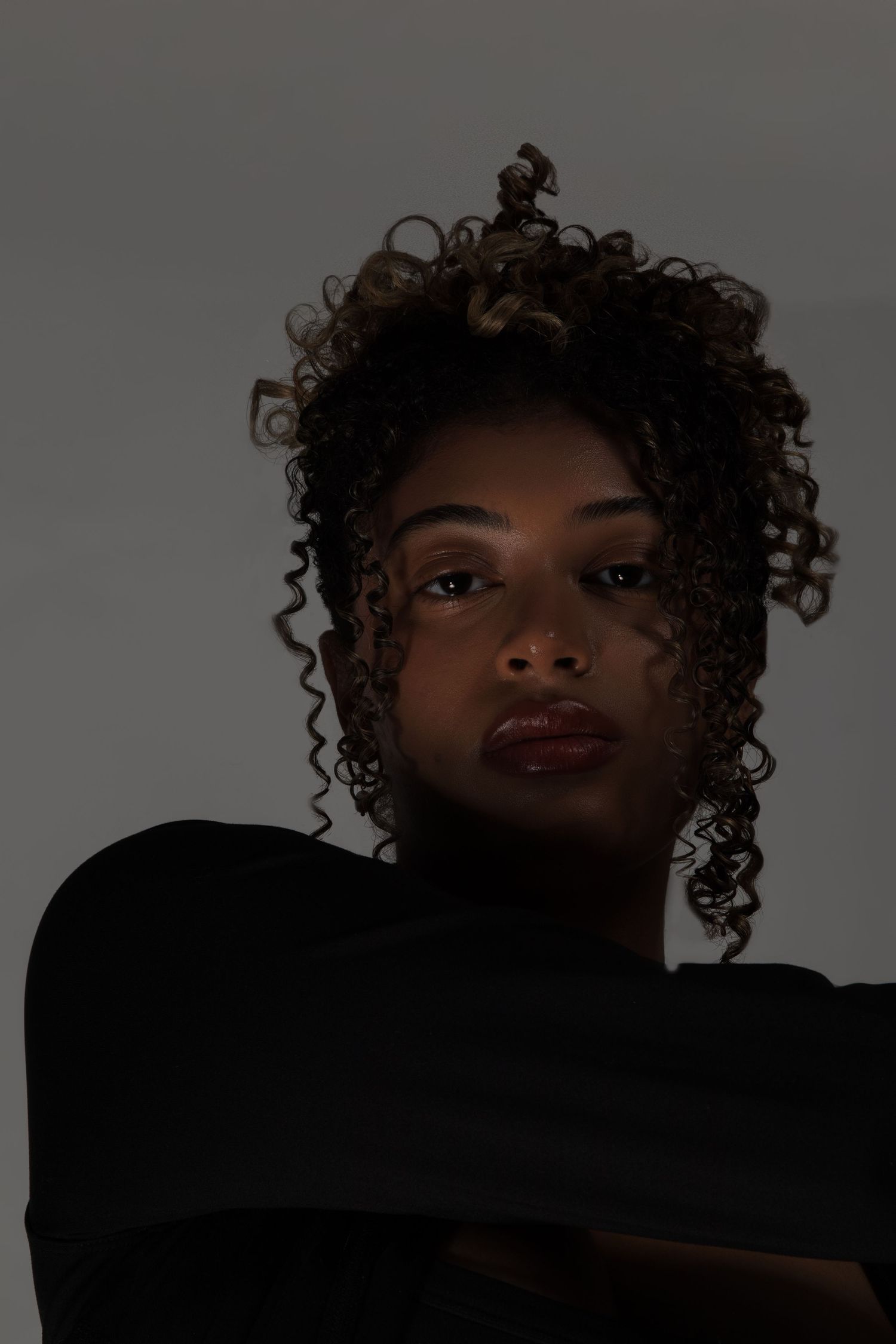 Close-up portrait of a young woman with natural curly hair wearing a black top in dramatic lighting.