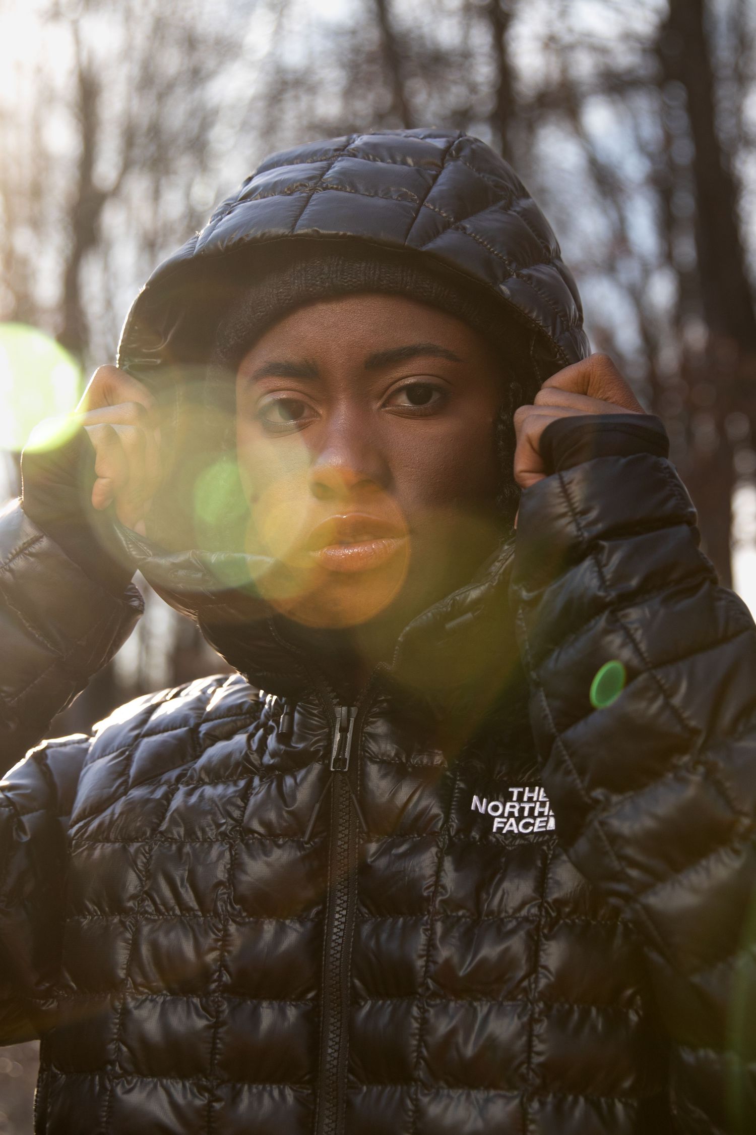 Woman in black North Face puffer jacket and beanie with sunlight lens flare in winter woods.