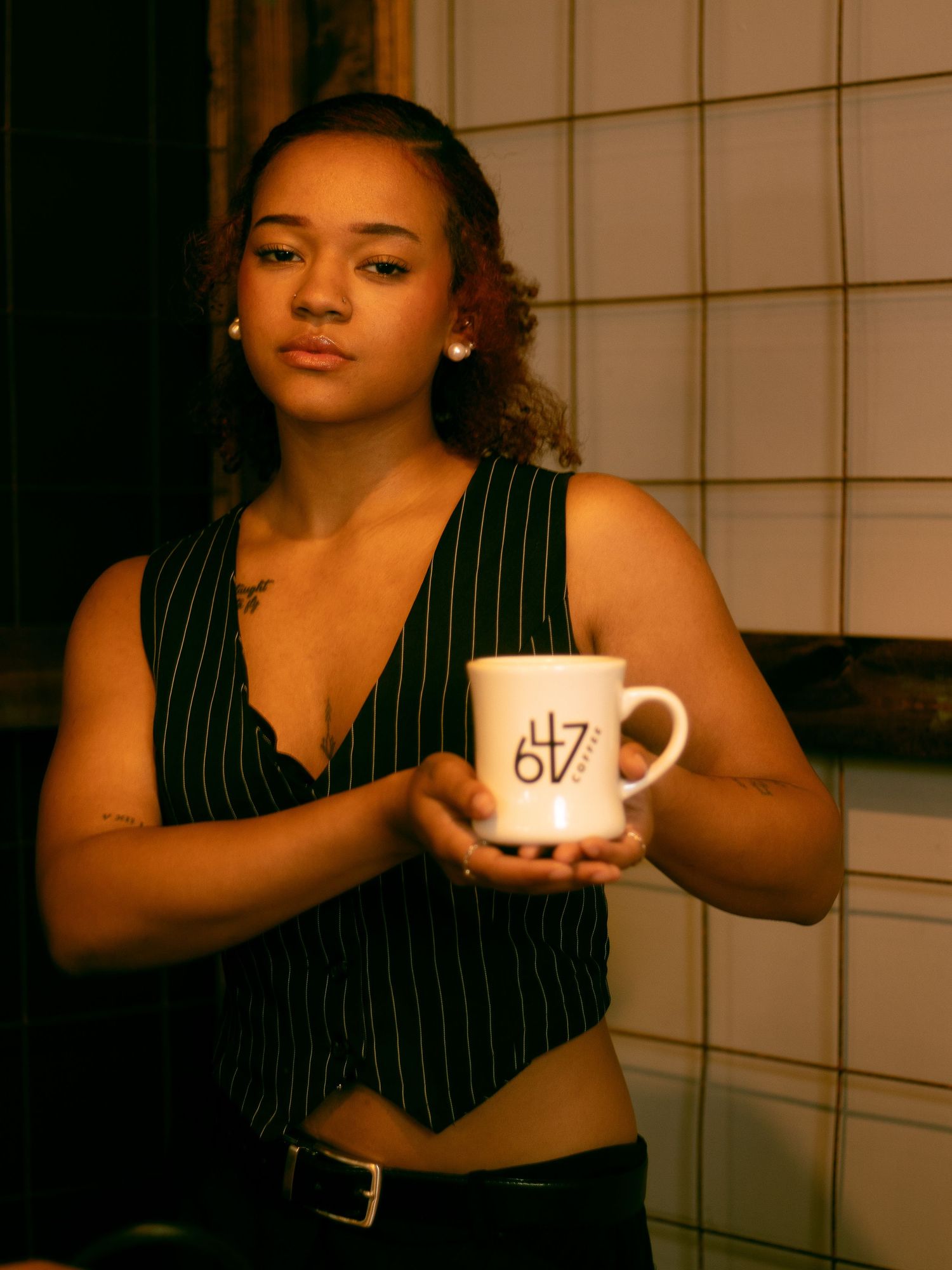 Young woman holding white branded mug wearing black pinstripe vest in cafe setting.