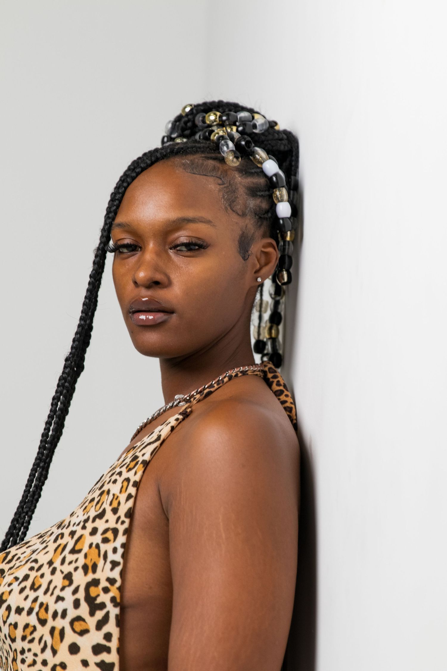 Black woman with beaded black braids leans against white wall wearing leopard print top and chain necklace.
