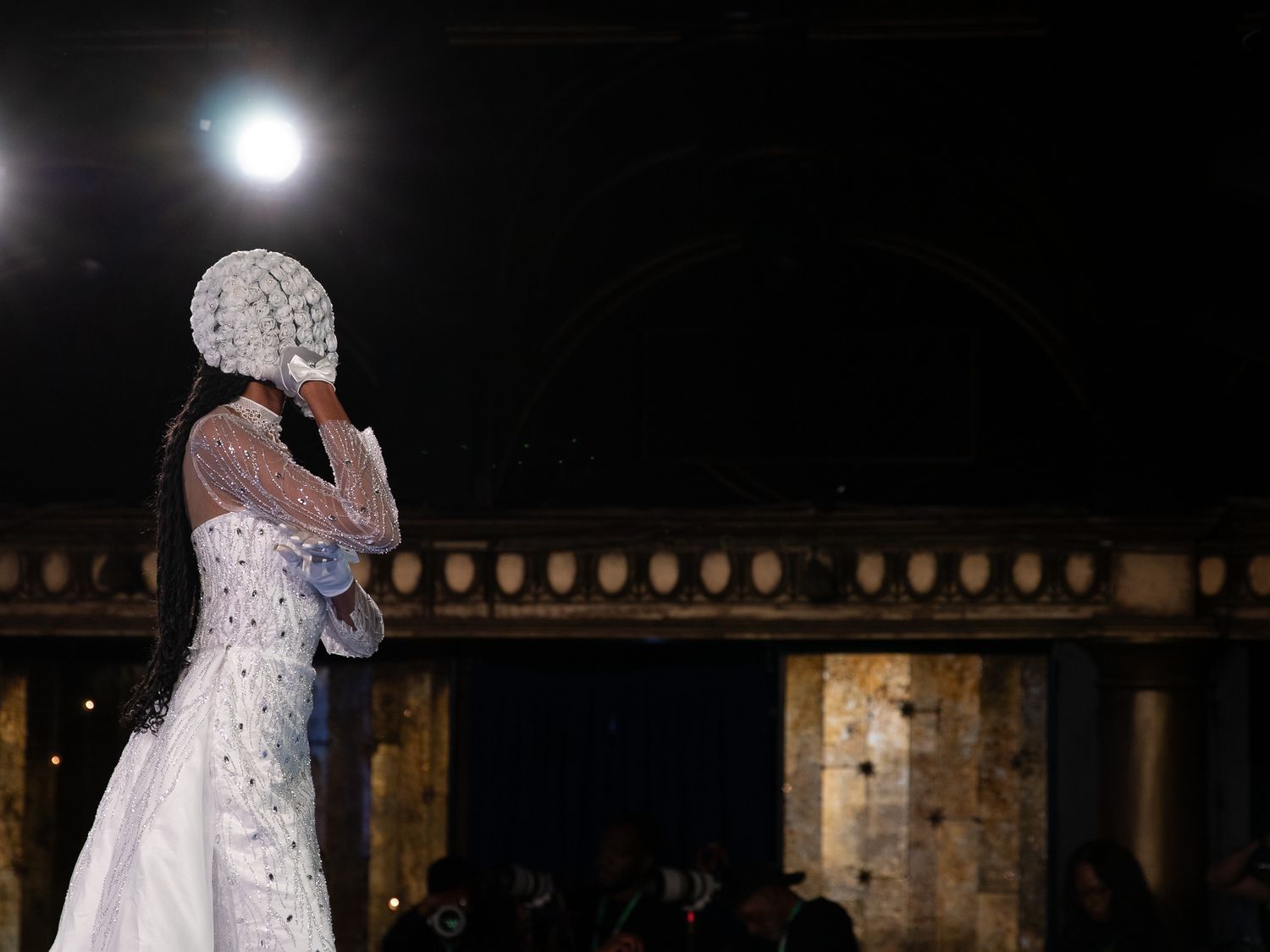 Model in elegant white bridal gown with ornate headpiece poses on runway at fashion show.