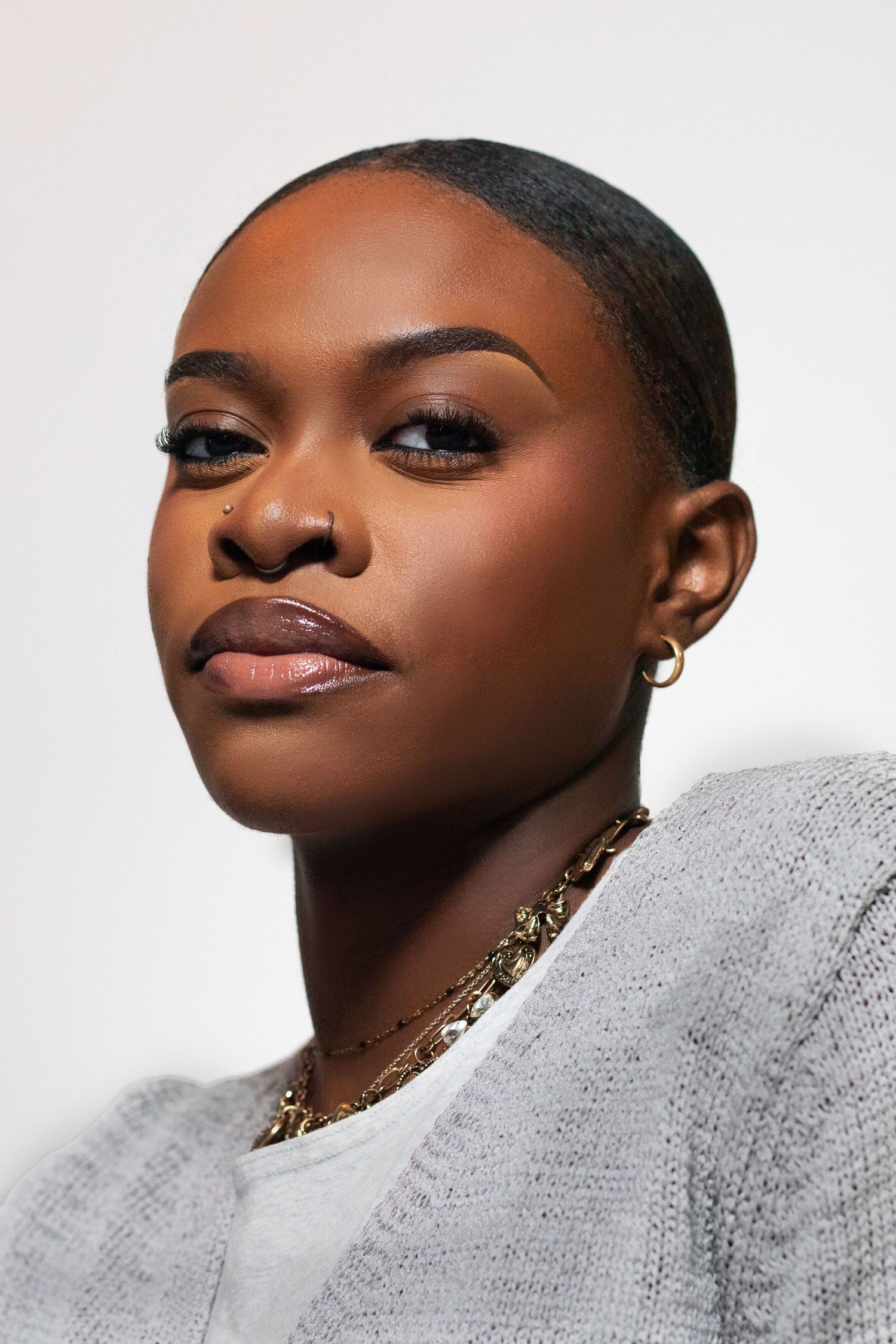 Confident young Black woman with shaved head, nose ring, gold earring and chain necklace in gray top.