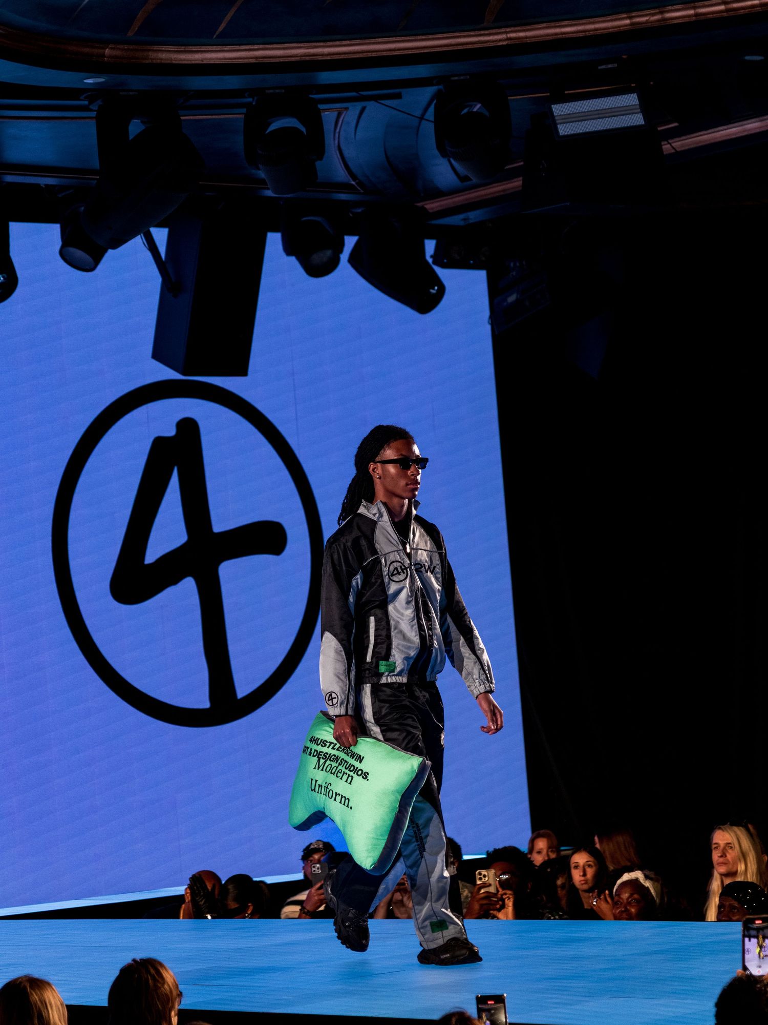 Male model walks runway in metallic jacket and baggy jeans carrying teal bag with number 4 logo backdrop.