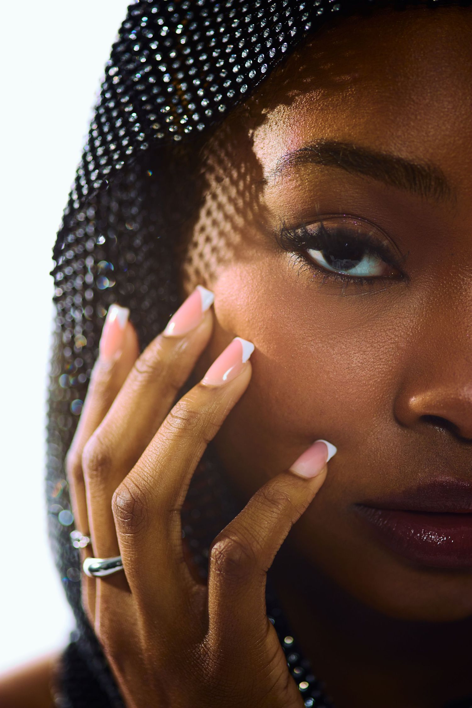 Close-up of a Black woman in a sequined black hood gently touching her cheek with manicured nails.