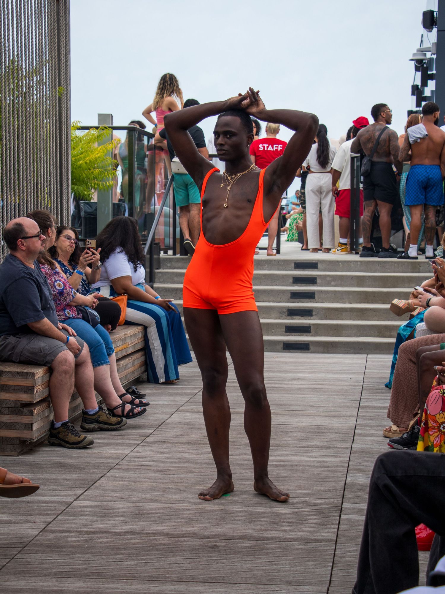 Male model in orange one-piece swimsuit poses with hands behind head on outdoor fashion runway.