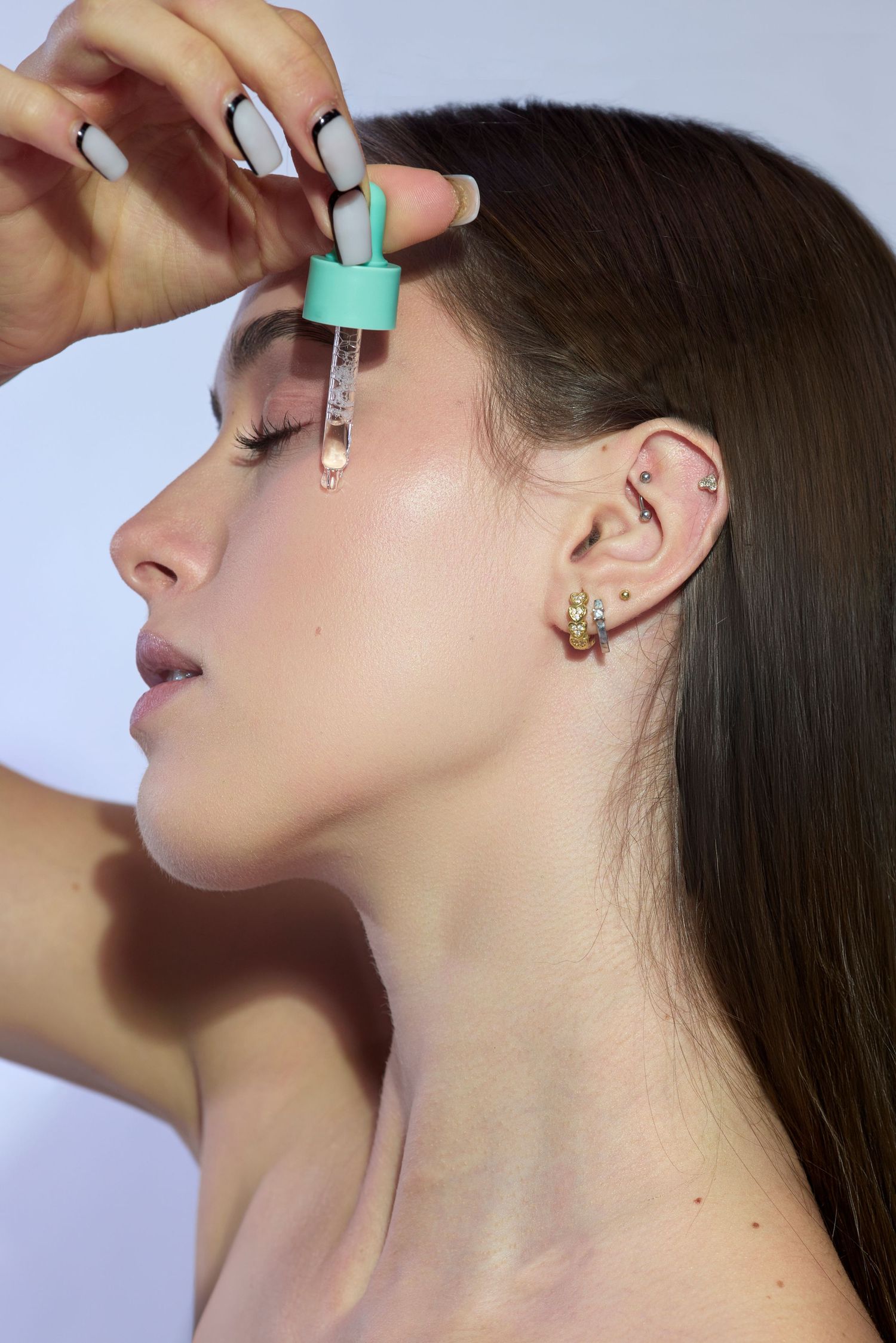 Young woman applying facial serum with dropper, showcasing skincare routine with gold earrings on light background.