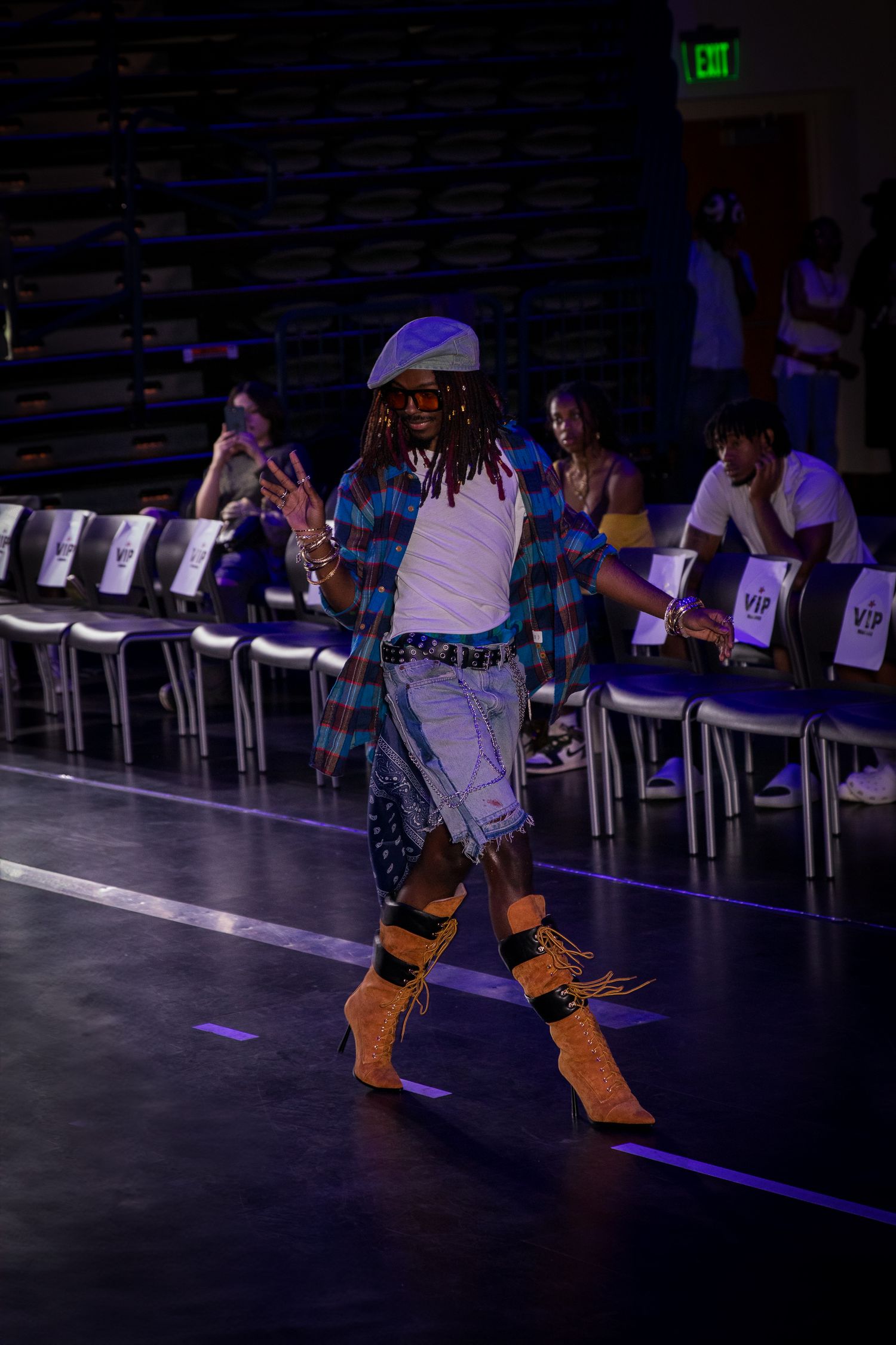 A model in a plaid shirt, denim shorts, and fringe boots walks an indoor fashion runway.