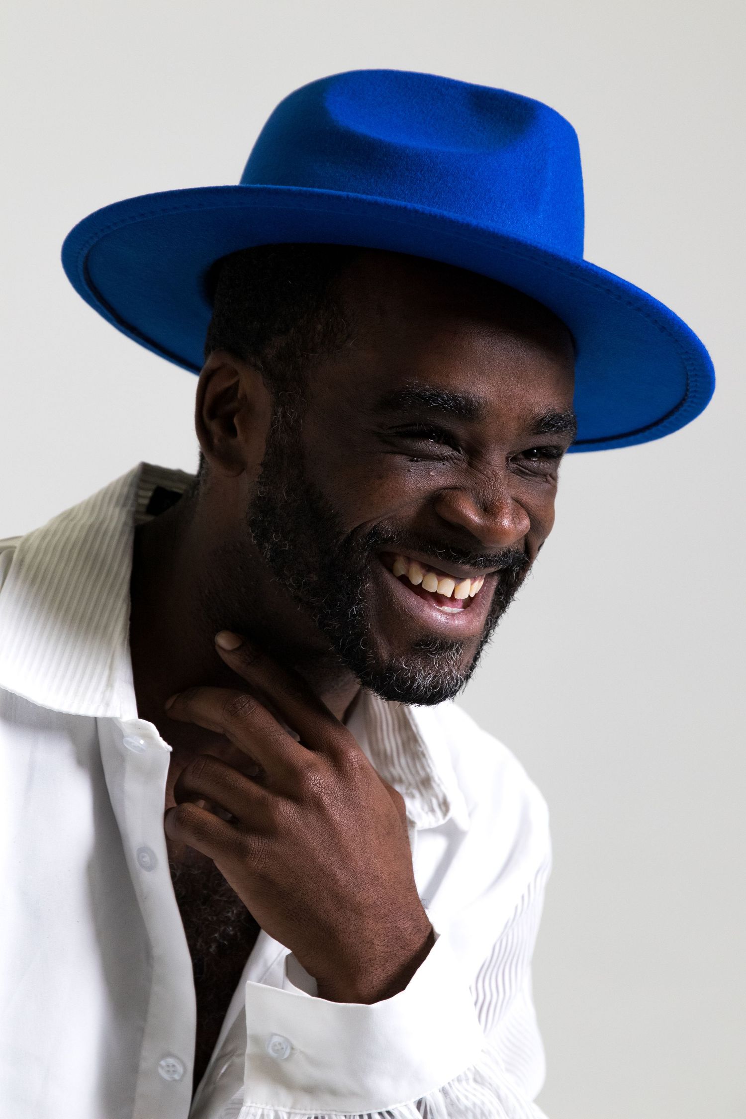 Smiling Black man wearing a bold blue wide-brim felt hat and white shirt against a light background.