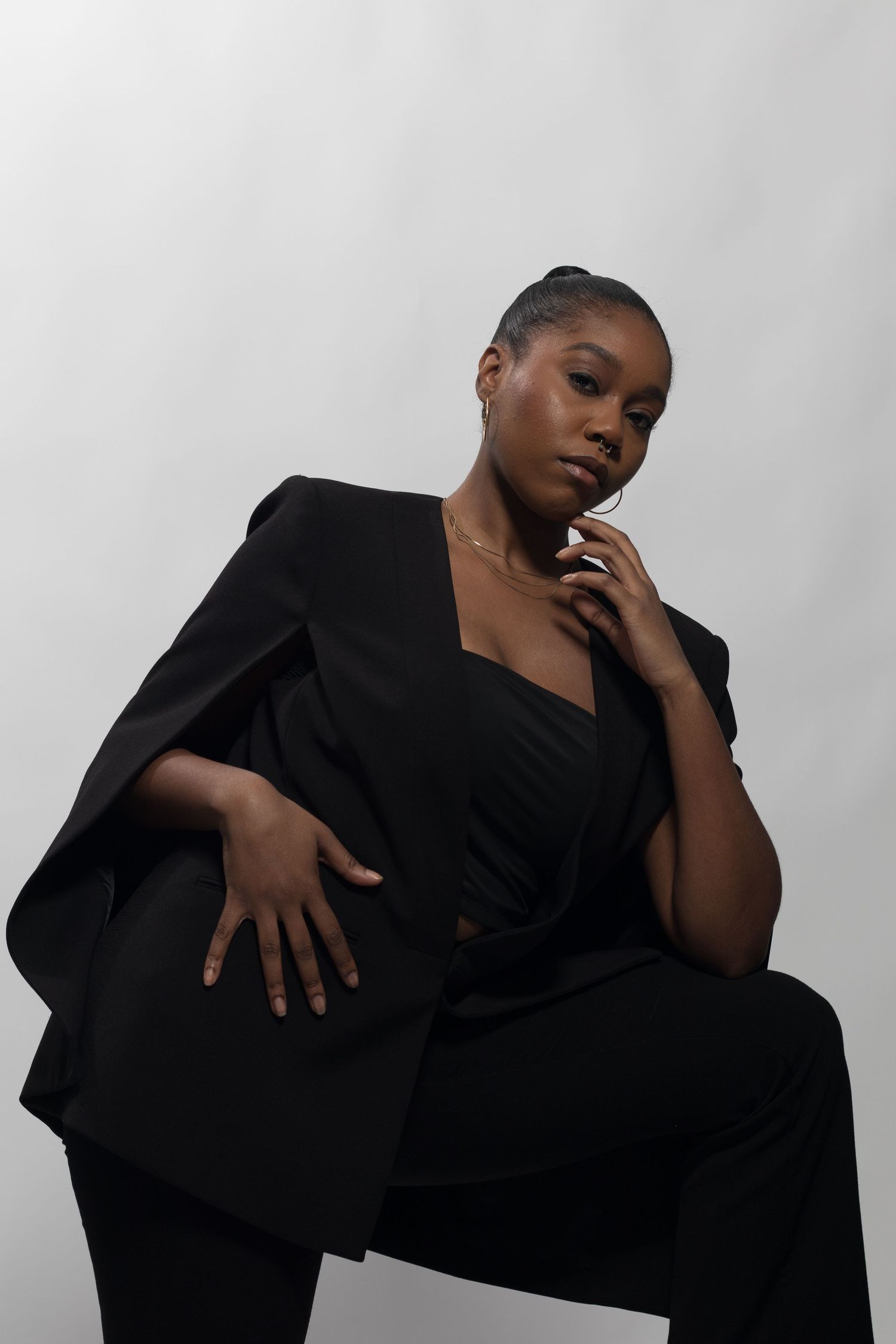 Black woman in black blazer and pants seated elegantly with hand near chin in studio.