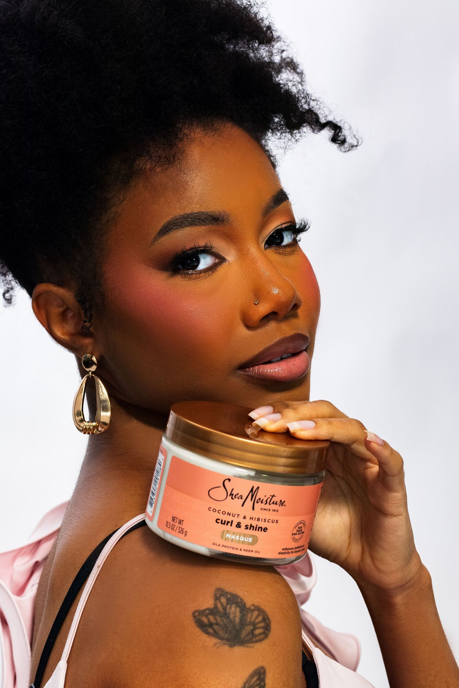 Woman with natural afro and butterfly tattoo holding SheaMoisture Curl & Shine mousse jar.
