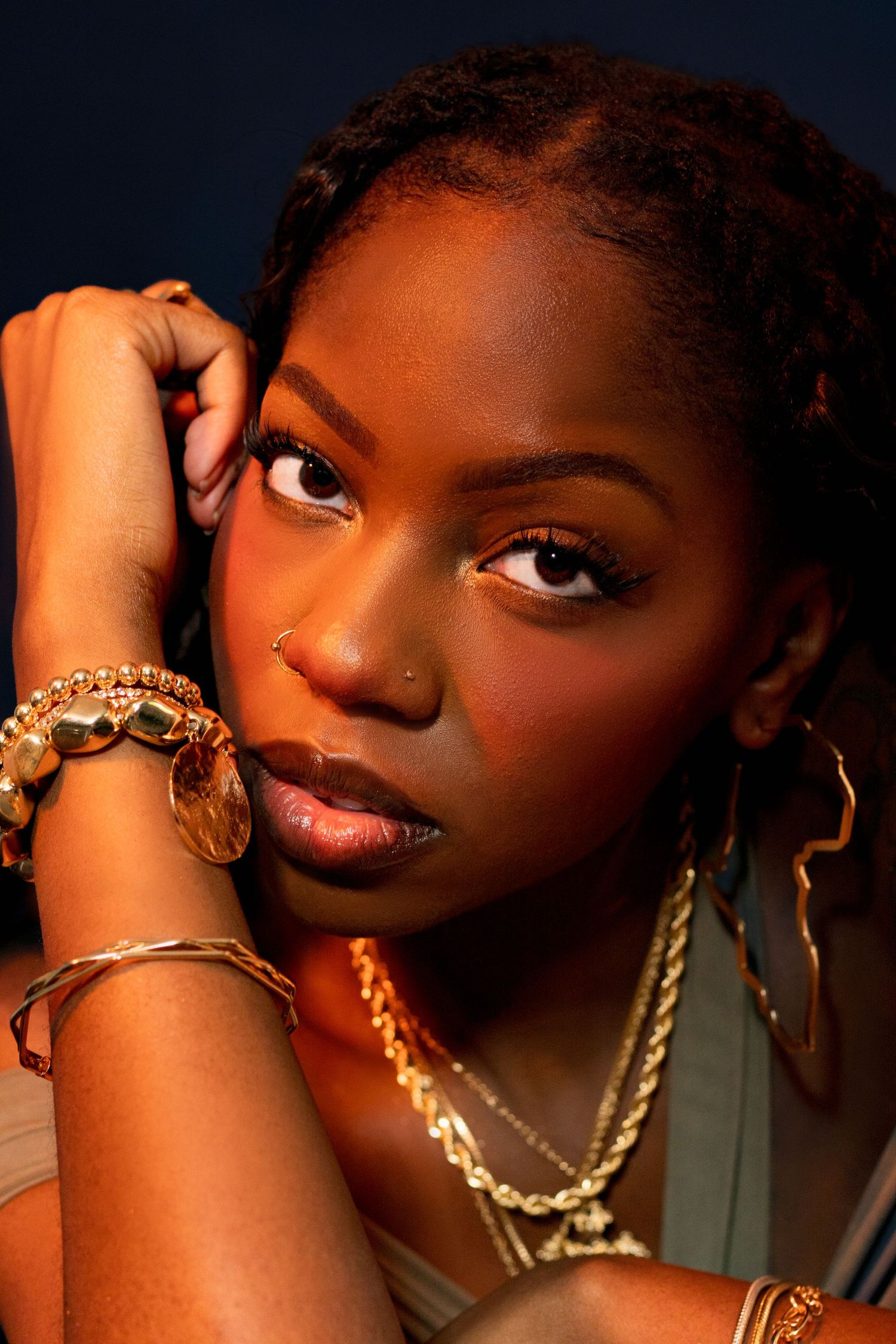 Close-up portrait of a Black woman with gold bracelets and necklaces resting head on hands in warm dramatic lighting.