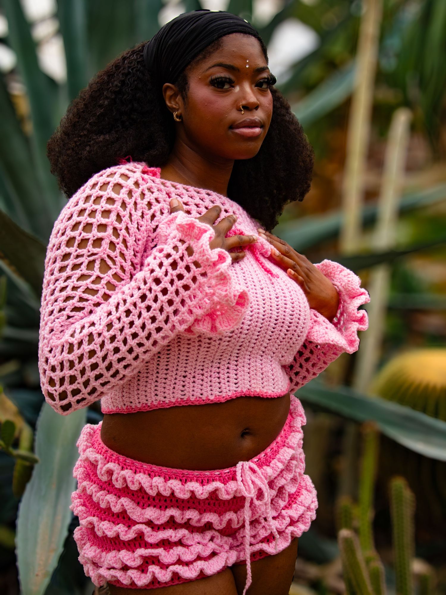 Black woman wearing pink crochet mesh top and ruffled skirt set with bindi accessory among desert plants.