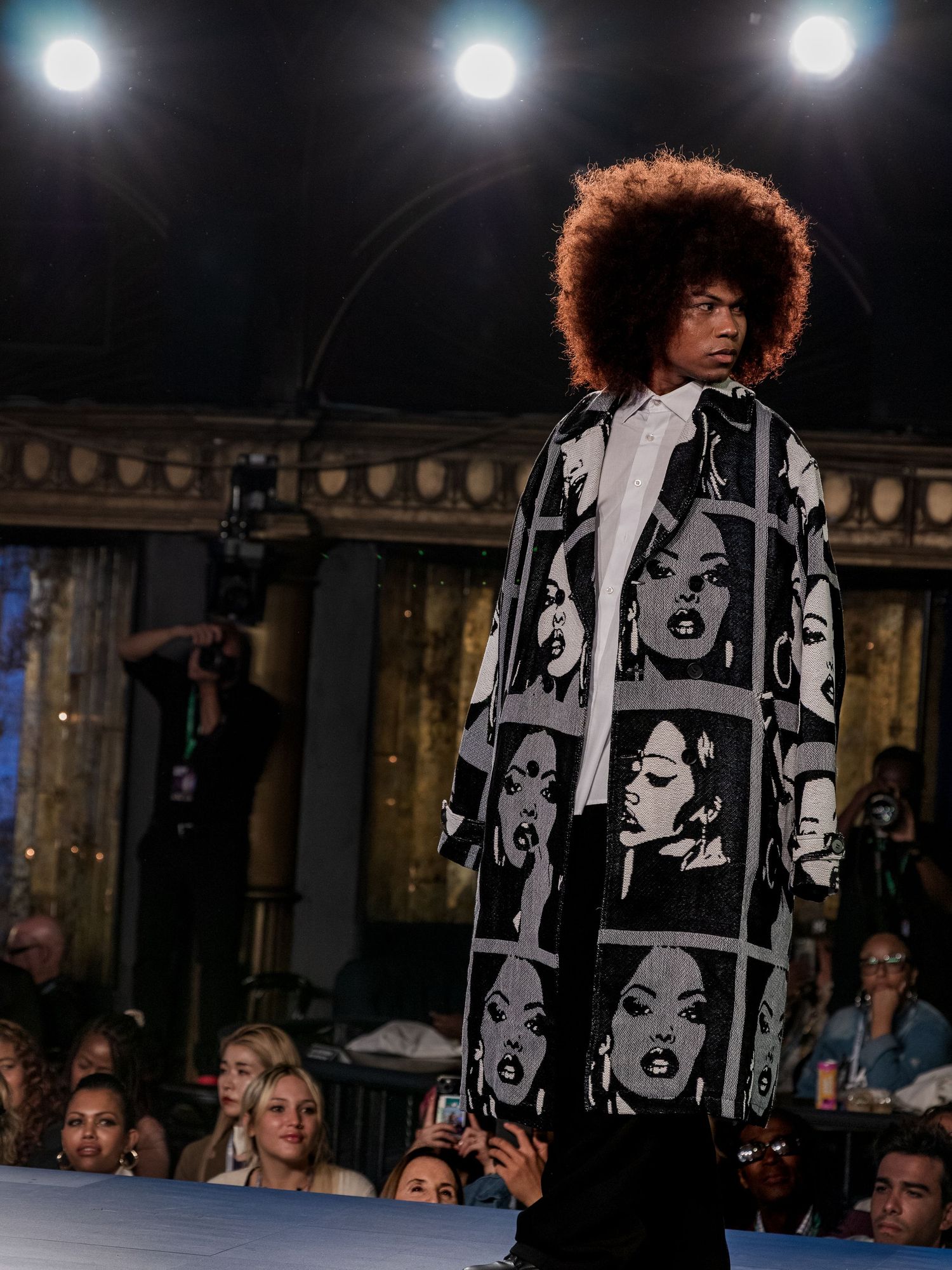 Male model with afro hairstyle wears bold black and white graphic face-print oversized coat on fashion runway.