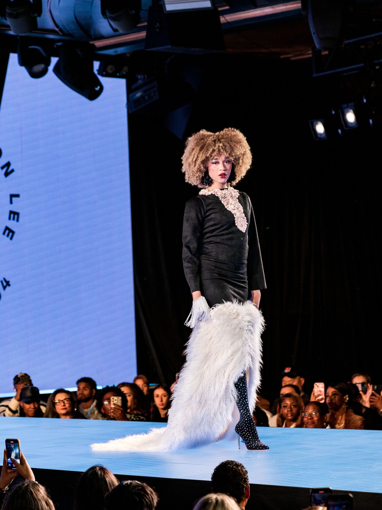 Model in black feather-trimmed gown walks runway with curly afro hairstyle at fashion show.
