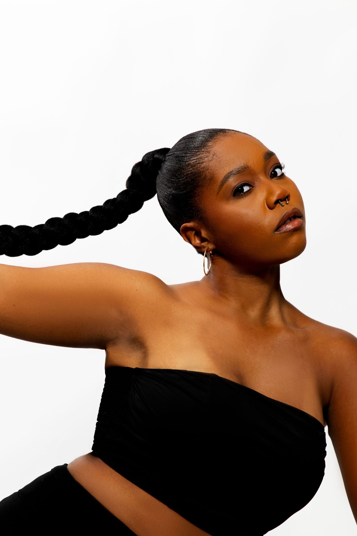 Black woman in black bandeau top with long braided ponytail posing confidently against white background.