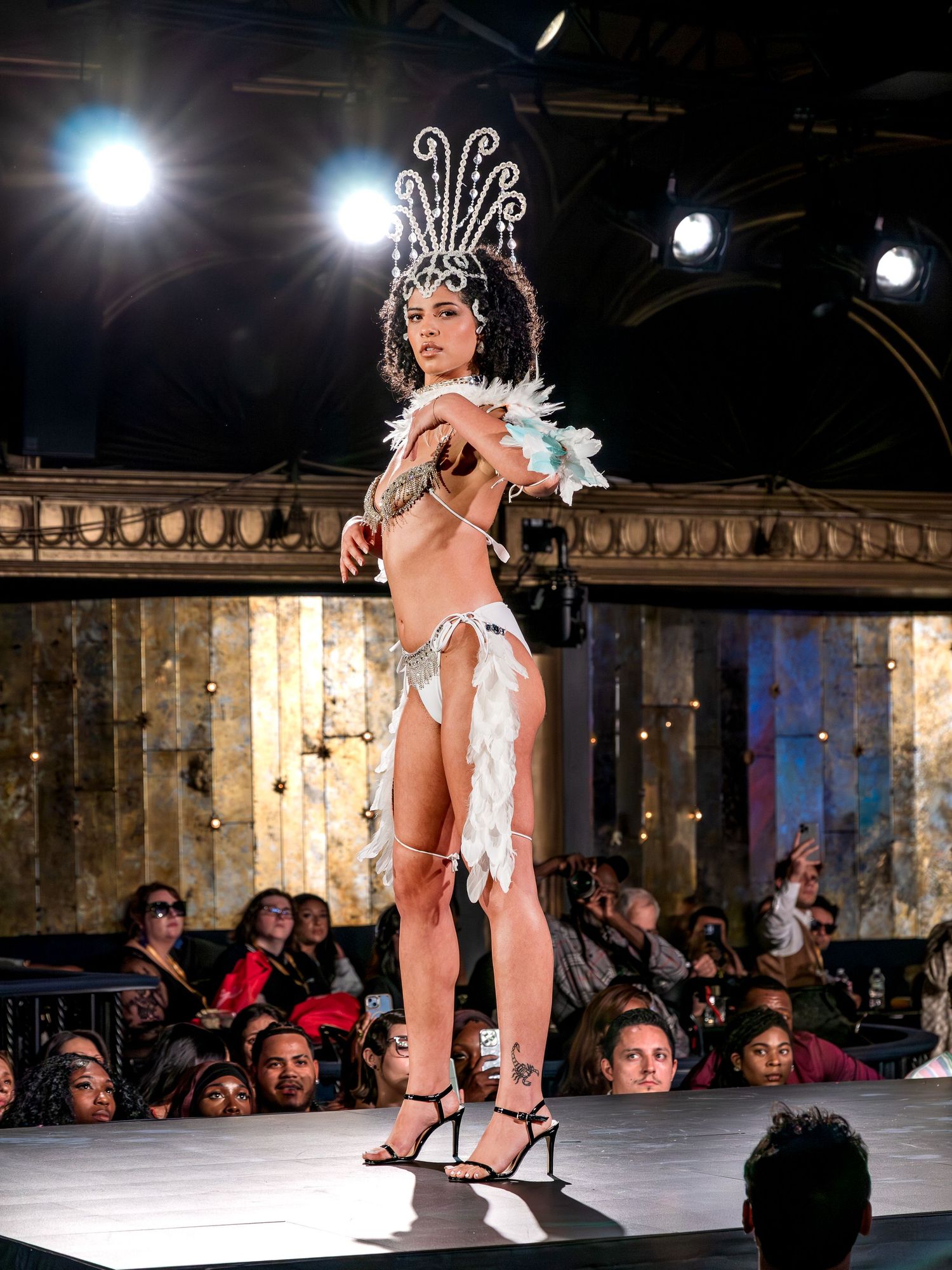Model on runway wearing white feathered bikini with elaborate crystal headdress before seated audience.