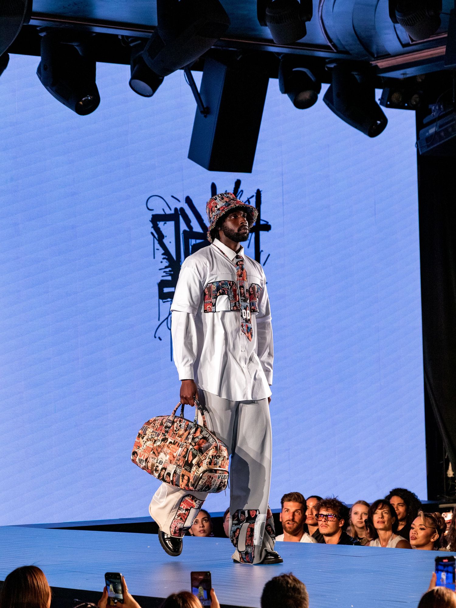 Male model walks runway in white outfit with graphic duffle bag and matching printed cap at fashion show.