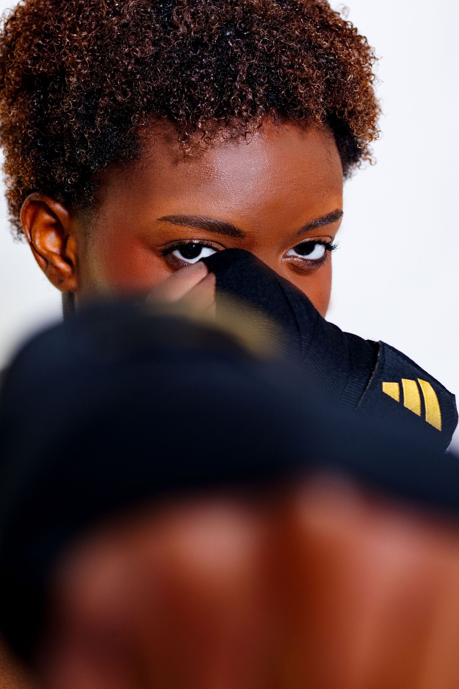 Intense close-up of a female boxer peering over a black and gold Adidas boxing glove with focused eyes.