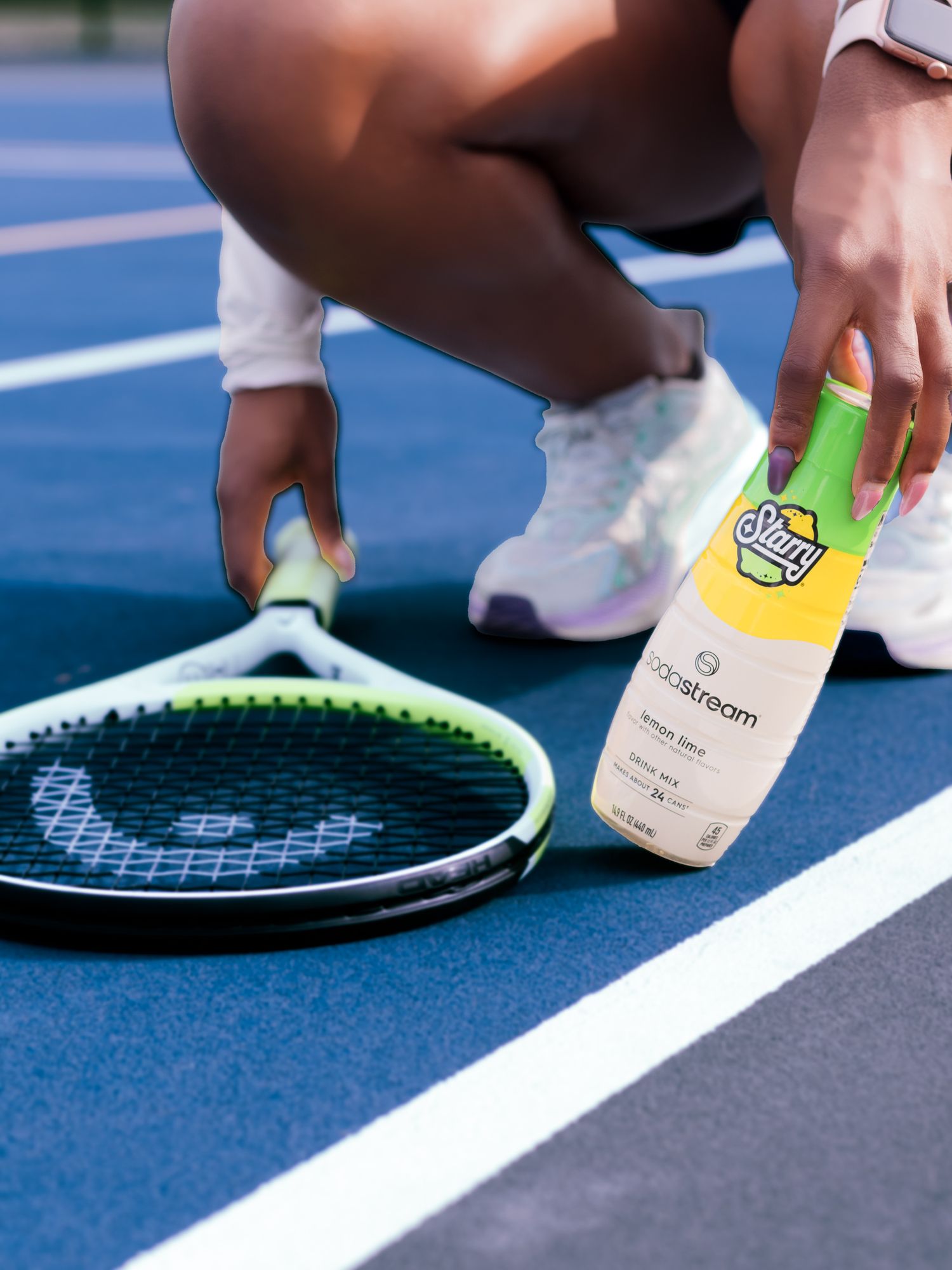Close-up of a person crouching on a blue tennis court holding a SodaStream bottle next to a tennis racket.
