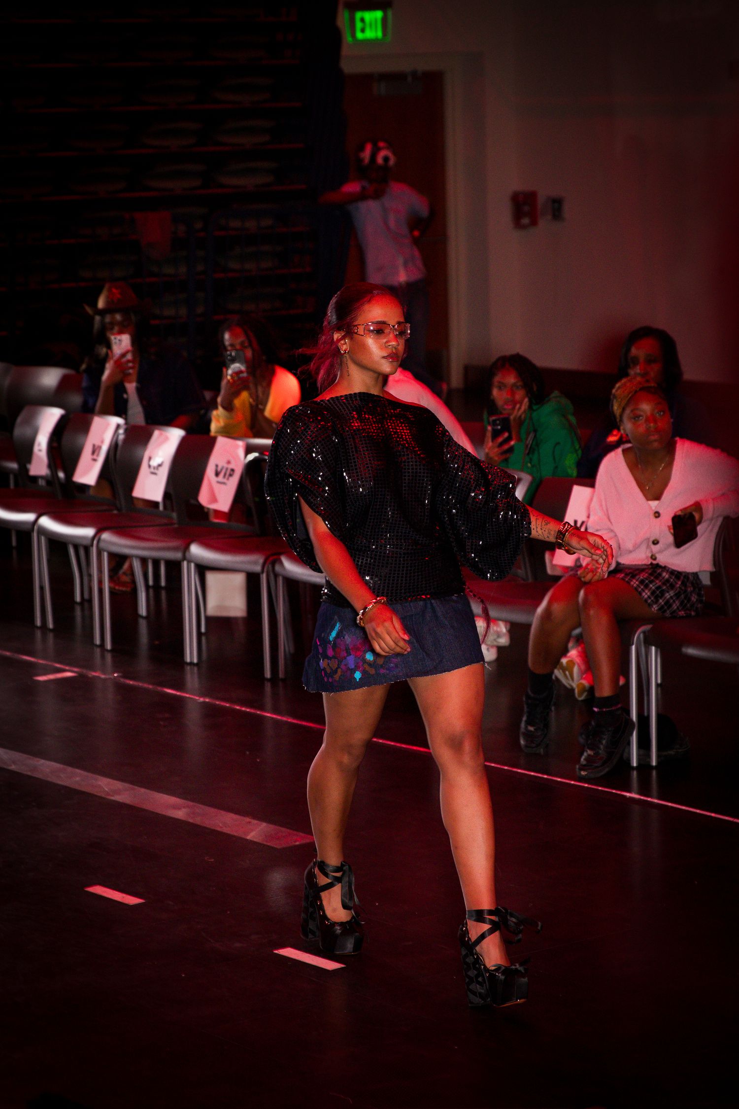 Model walks runway in sequin black jacket and holographic mini skirt under red lighting at fashion show.