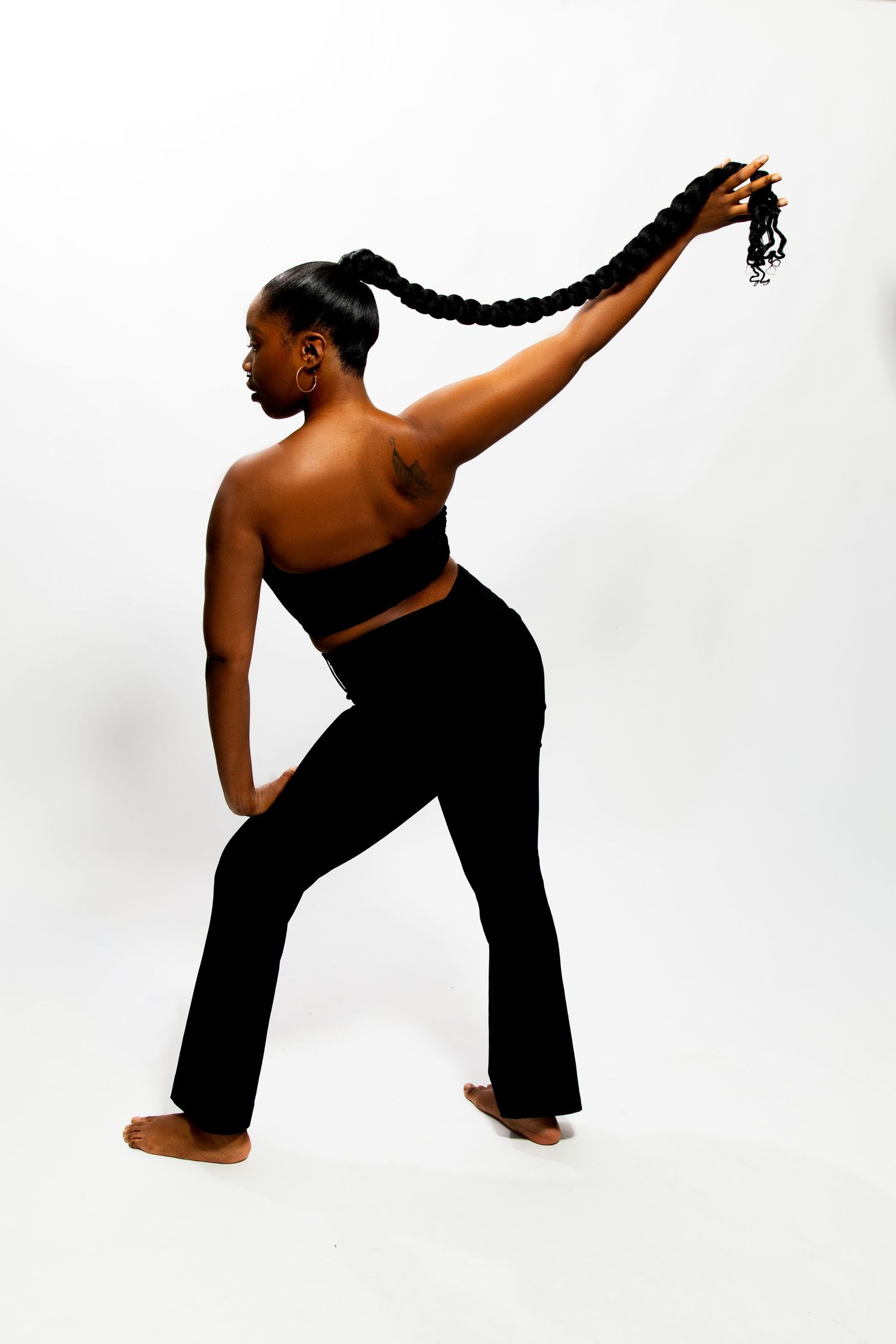 Black woman in black crop top and flared pants holding braided ponytail overhead from behind.
