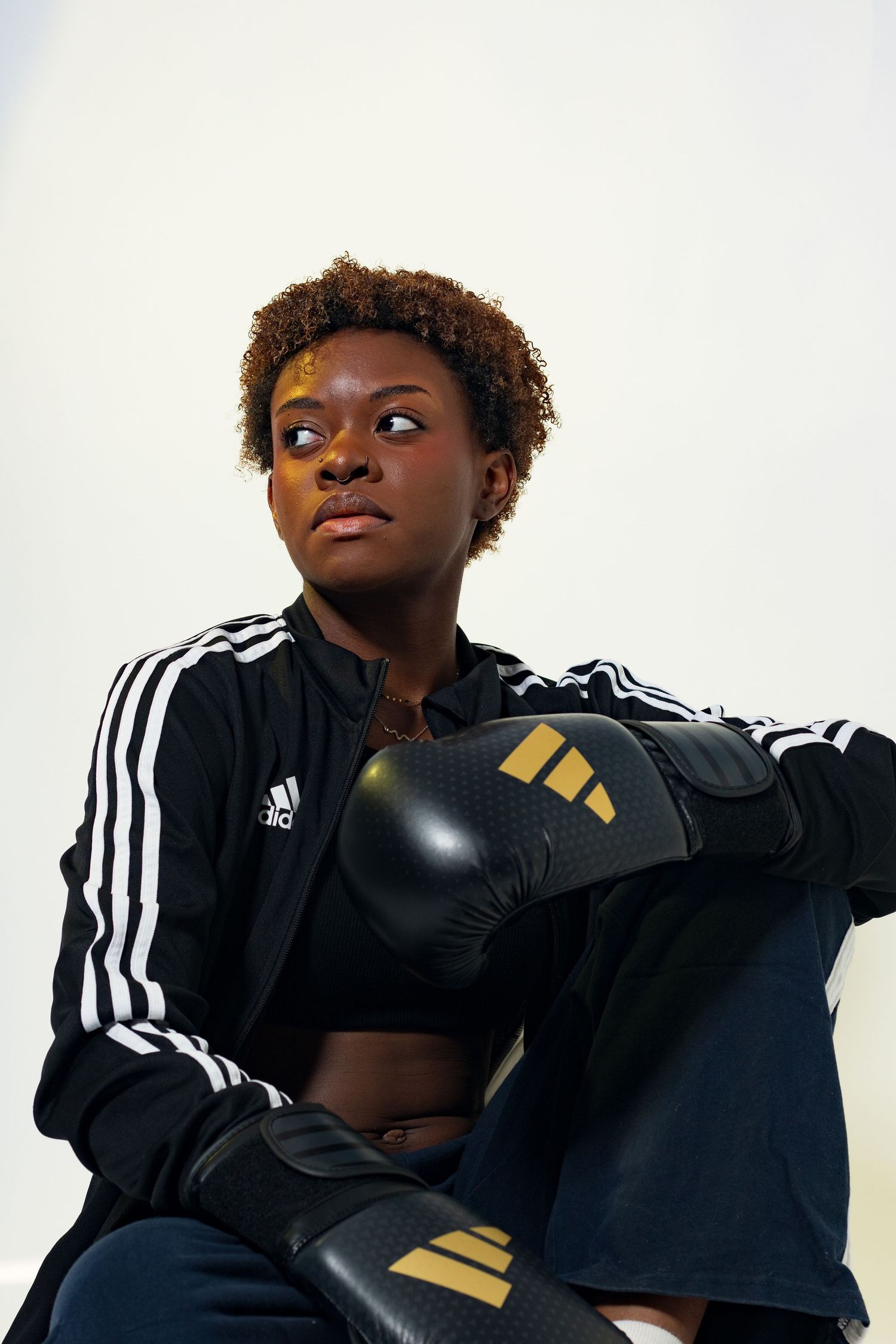 Young Black female boxer in Adidas tracksuit sitting relaxed with black and gold boxing gloves resting.