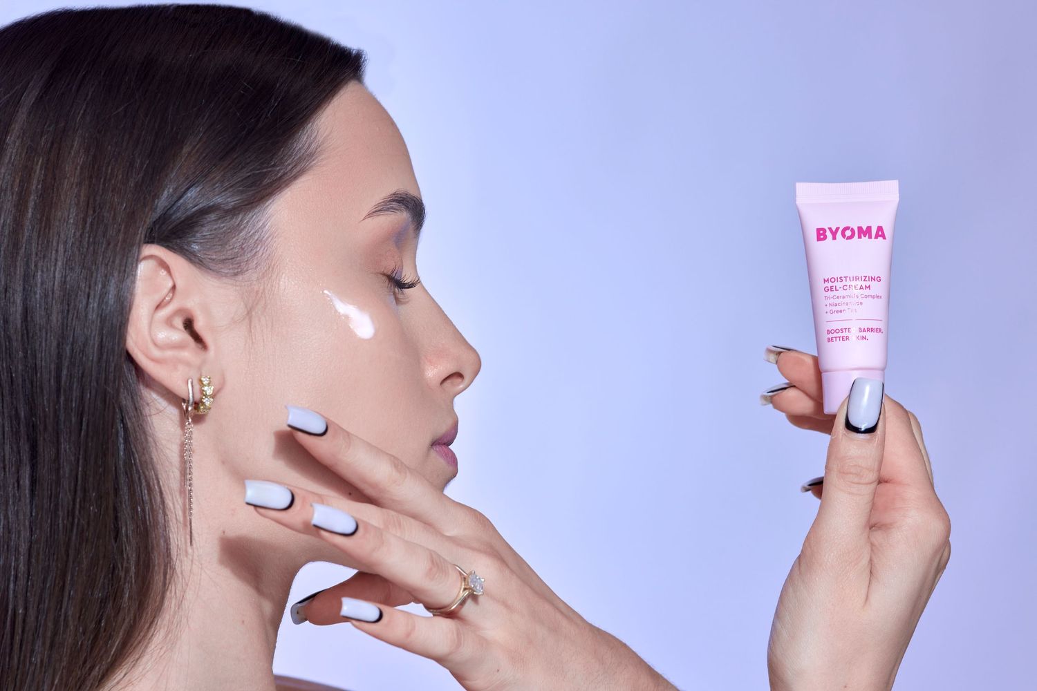 Woman applying BYOMA pink skincare cream tube to her cheek against a soft purple background.