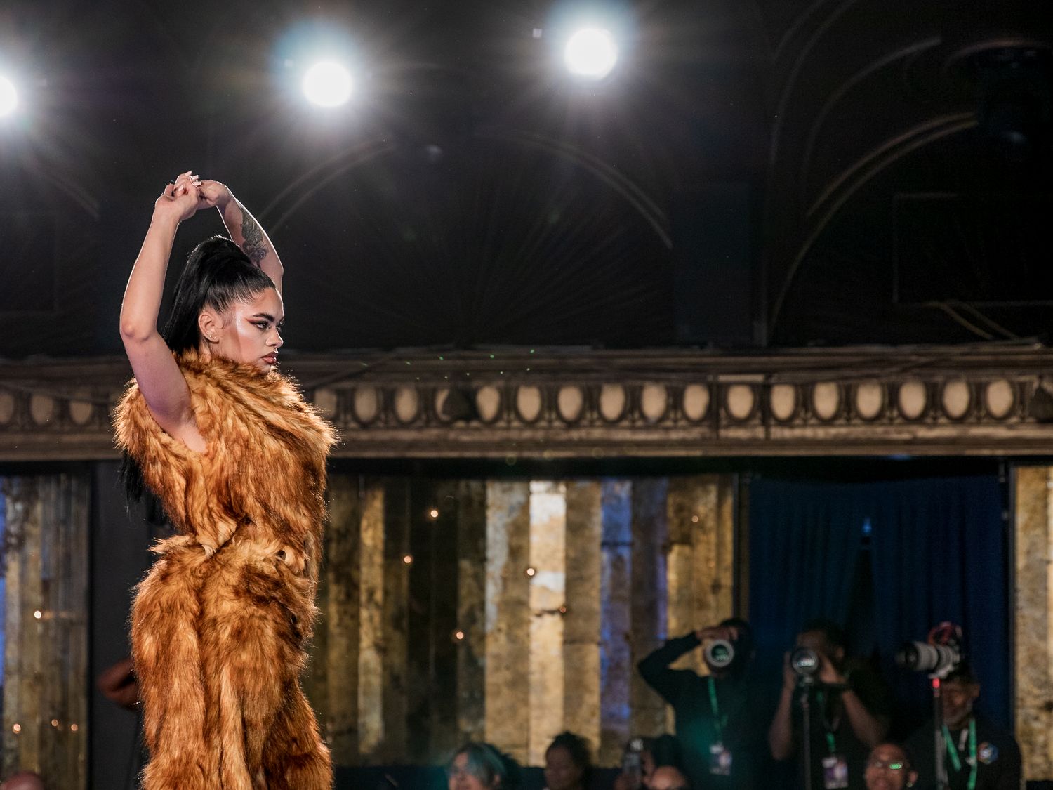 Model strikes dramatic pose in textured golden fur dress on runway at fashion show.