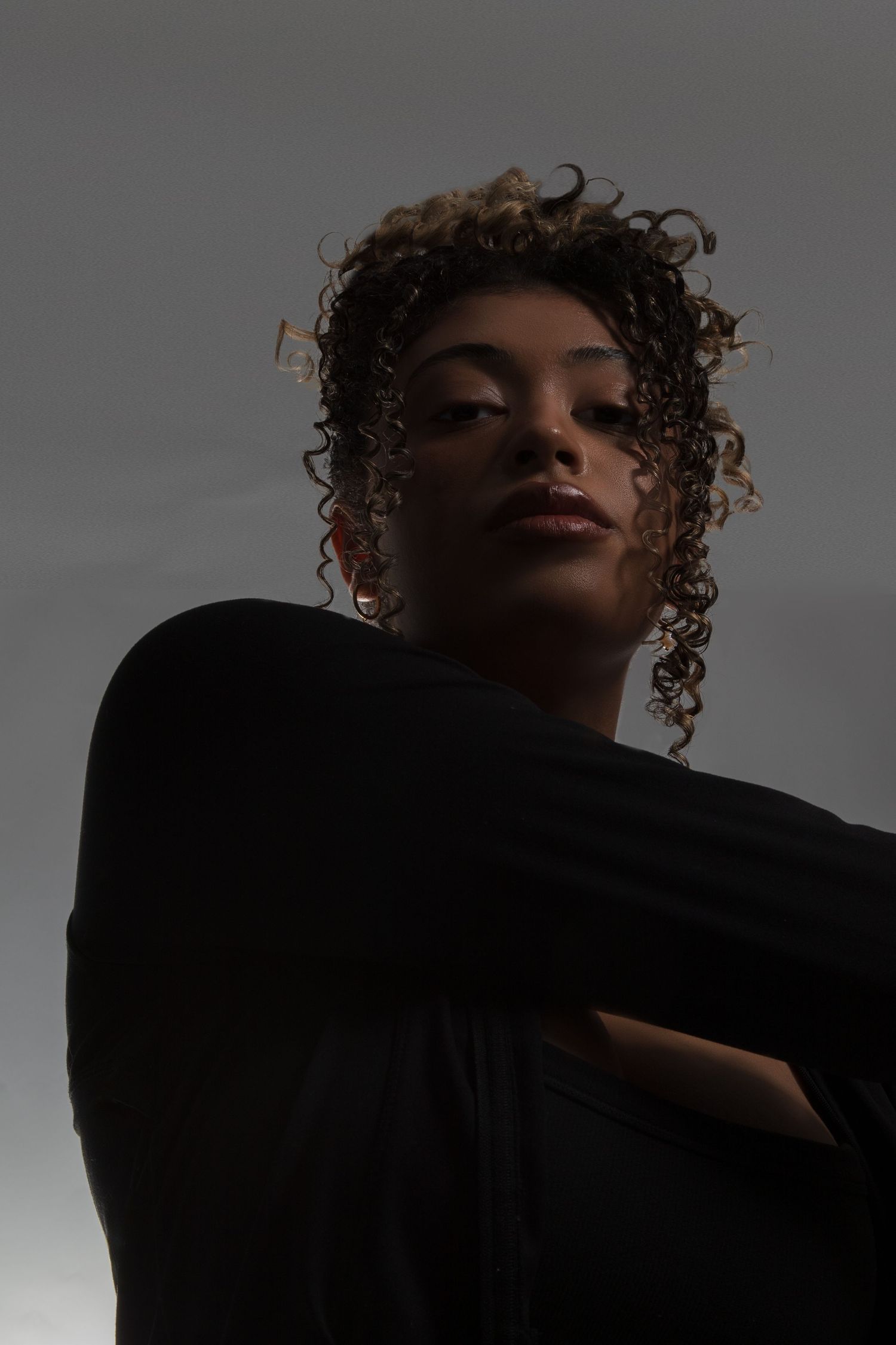 A young woman with curly hair poses confidently in a black outfit against a dark gray background.