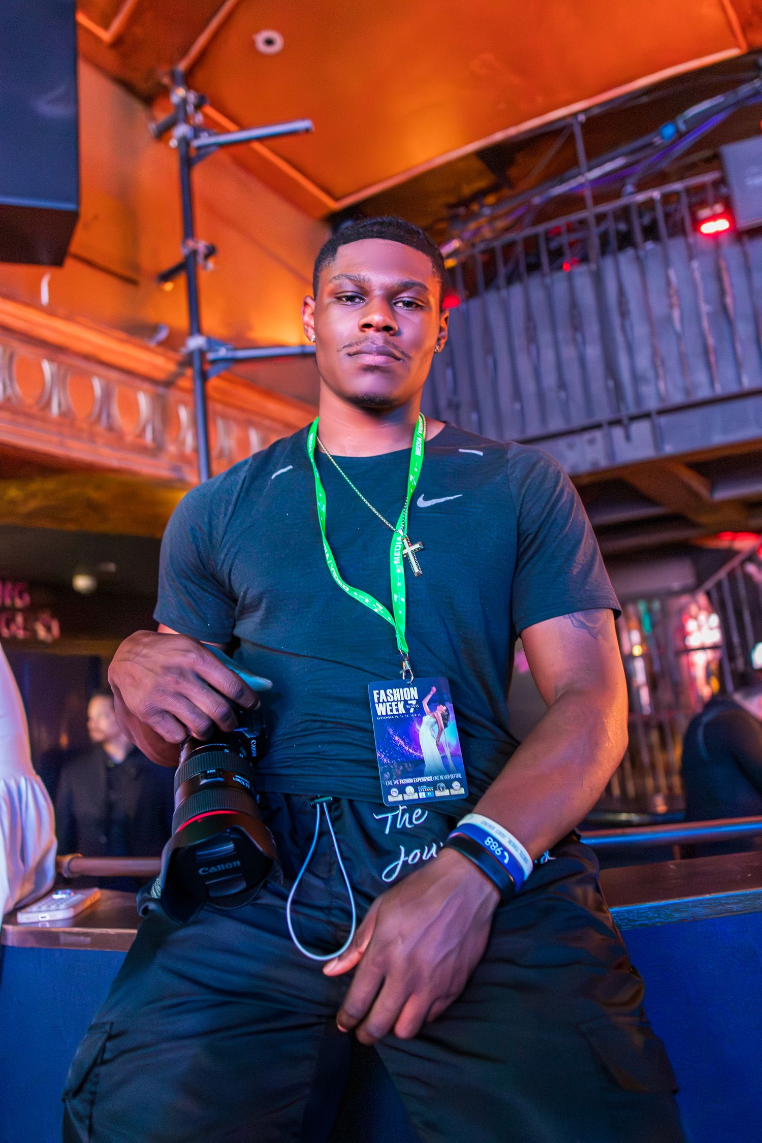 Young Black man holds camera while wearing Fashion Week badge and Nike shirt at venue.