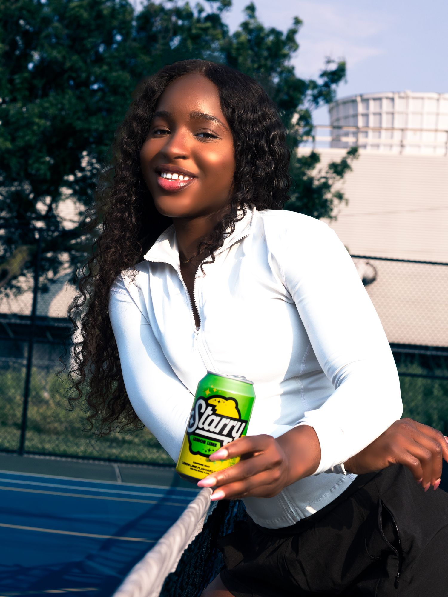 Smiling young woman holding a Starry lemon-lime soda can while leaning on a tennis net outdoors.