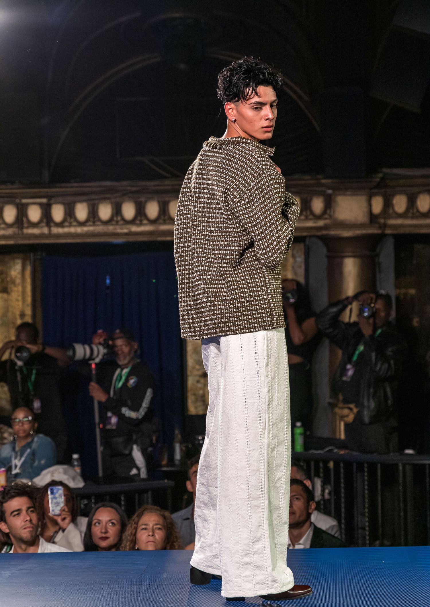 Male model showcasing patterned knit jacket and white wide-leg pants on a fashion runway at night.