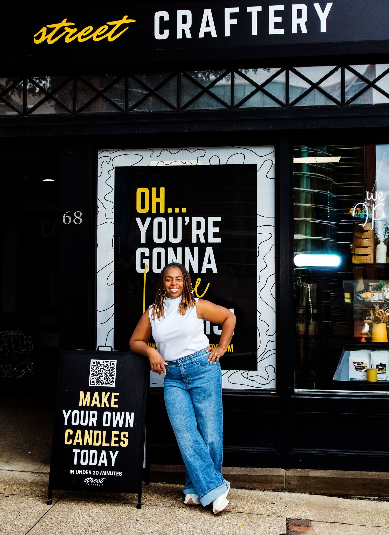 Smiling woman in white tank top posing outside Street Craftery shop with bold yellow window graphics.
