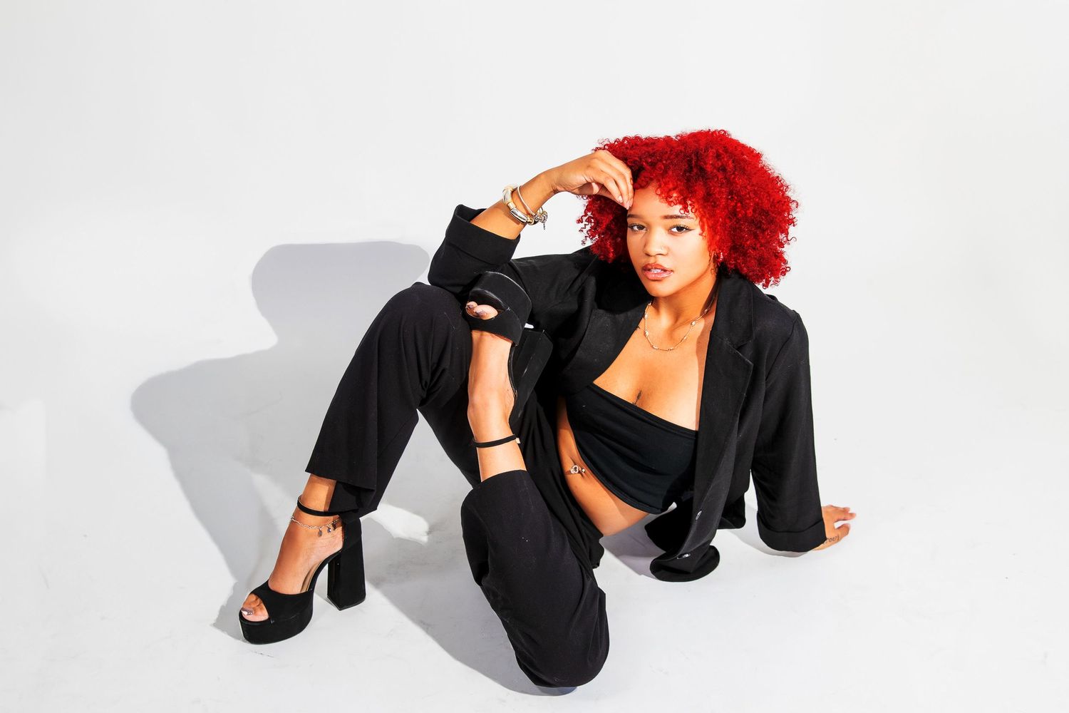 Black woman with vibrant red afro in black blazer and pants posing on white studio floor.