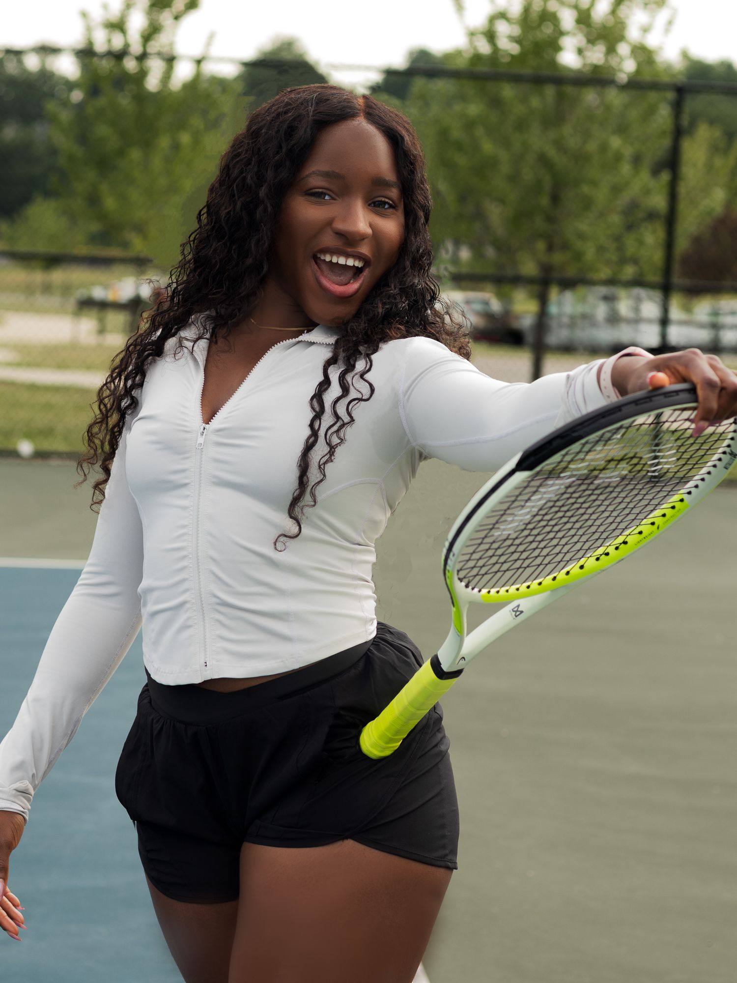Smiling young woman in white athletic top and black shorts holding a tennis racket on an outdoor court.