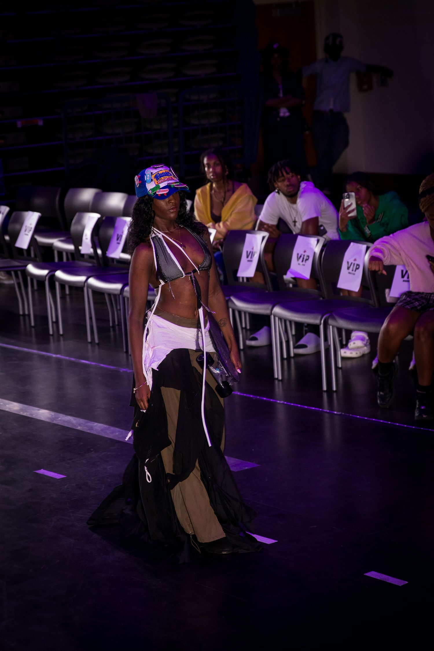 Model walks runway in avant-garde black and white outfit with colorful headpiece at VIP fashion show.