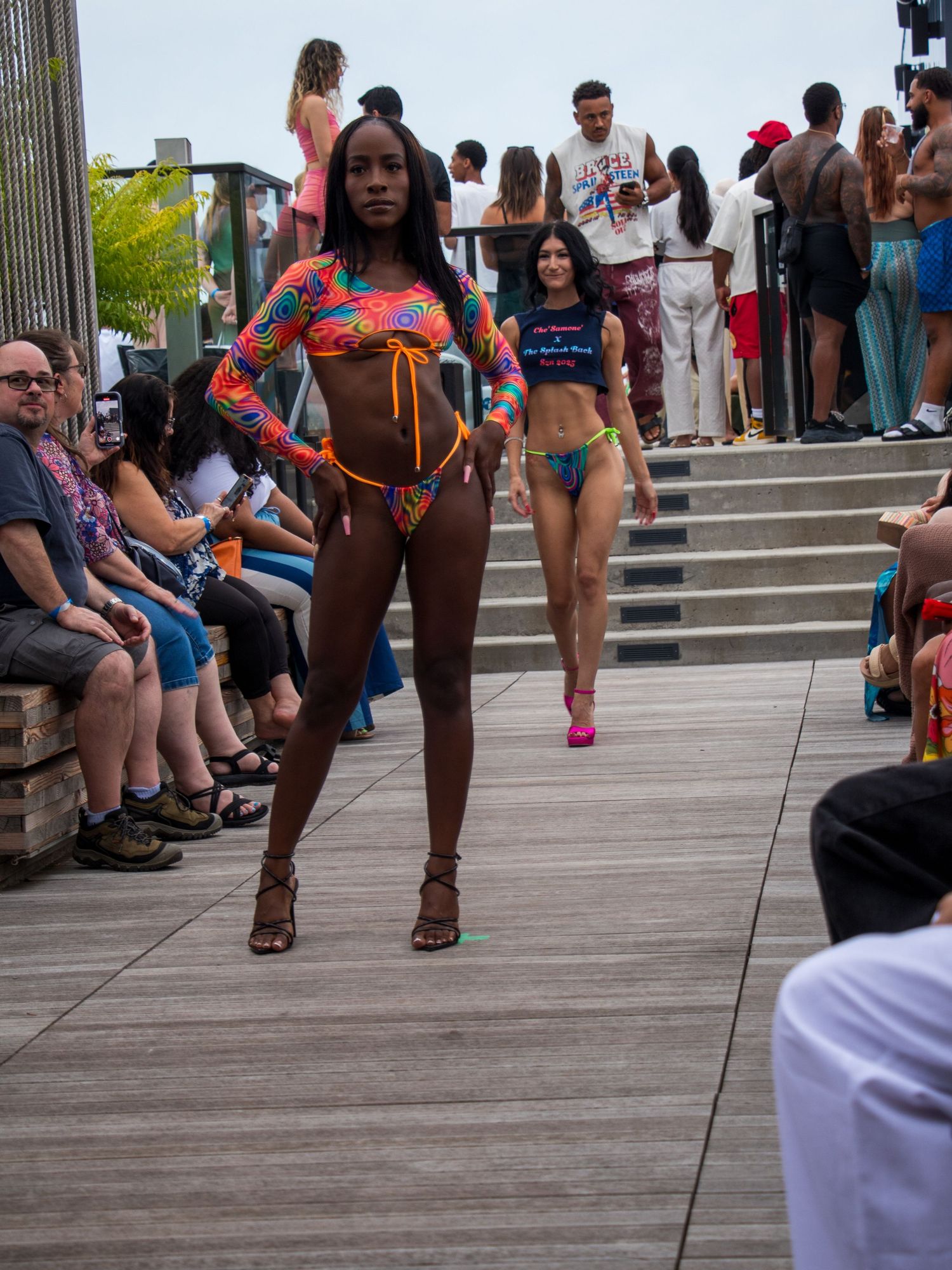 Models walk an outdoor runway in colorful swimwear during a daytime beach fashion show.