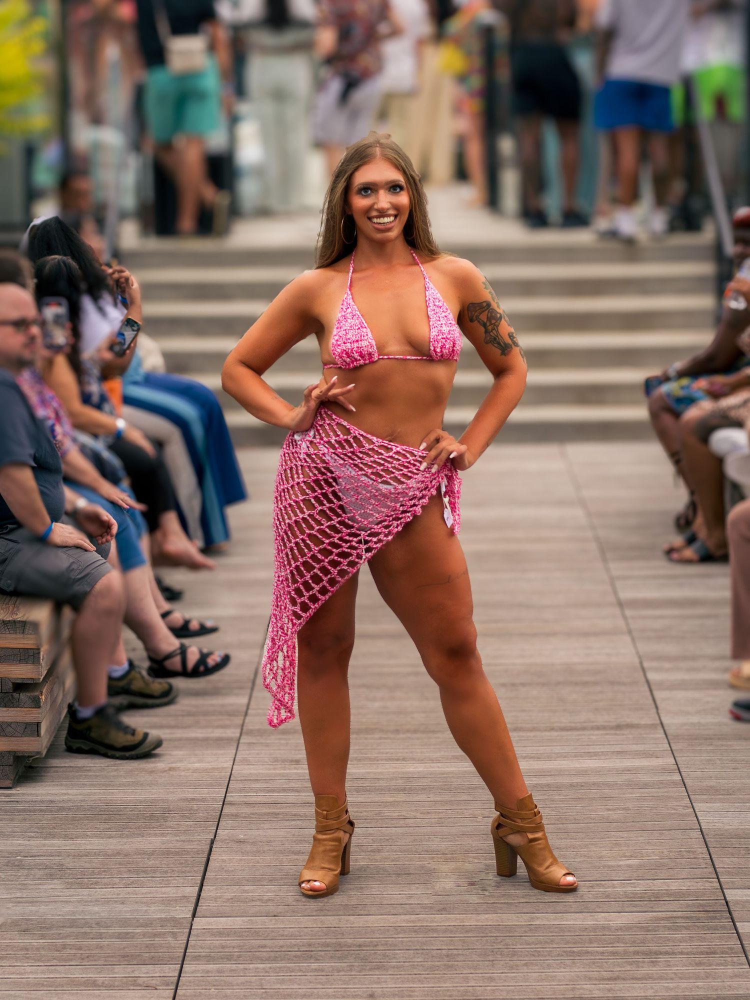 Woman in pink crochet bikini and net wrap skirt poses on outdoor boardwalk fashion runway.