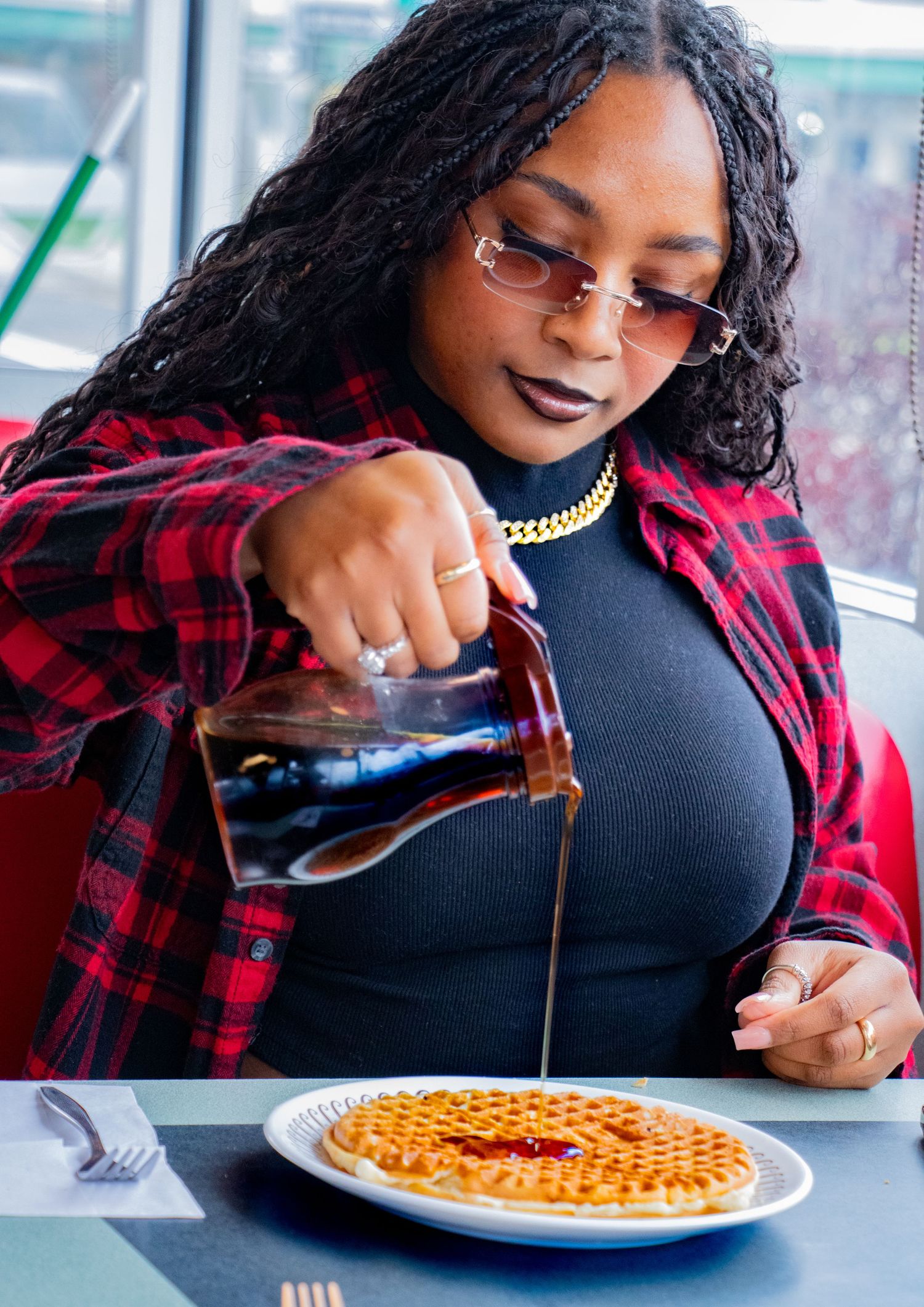Woman in plaid shirt pouring maple syrup on a golden waffle at a restaurant table.