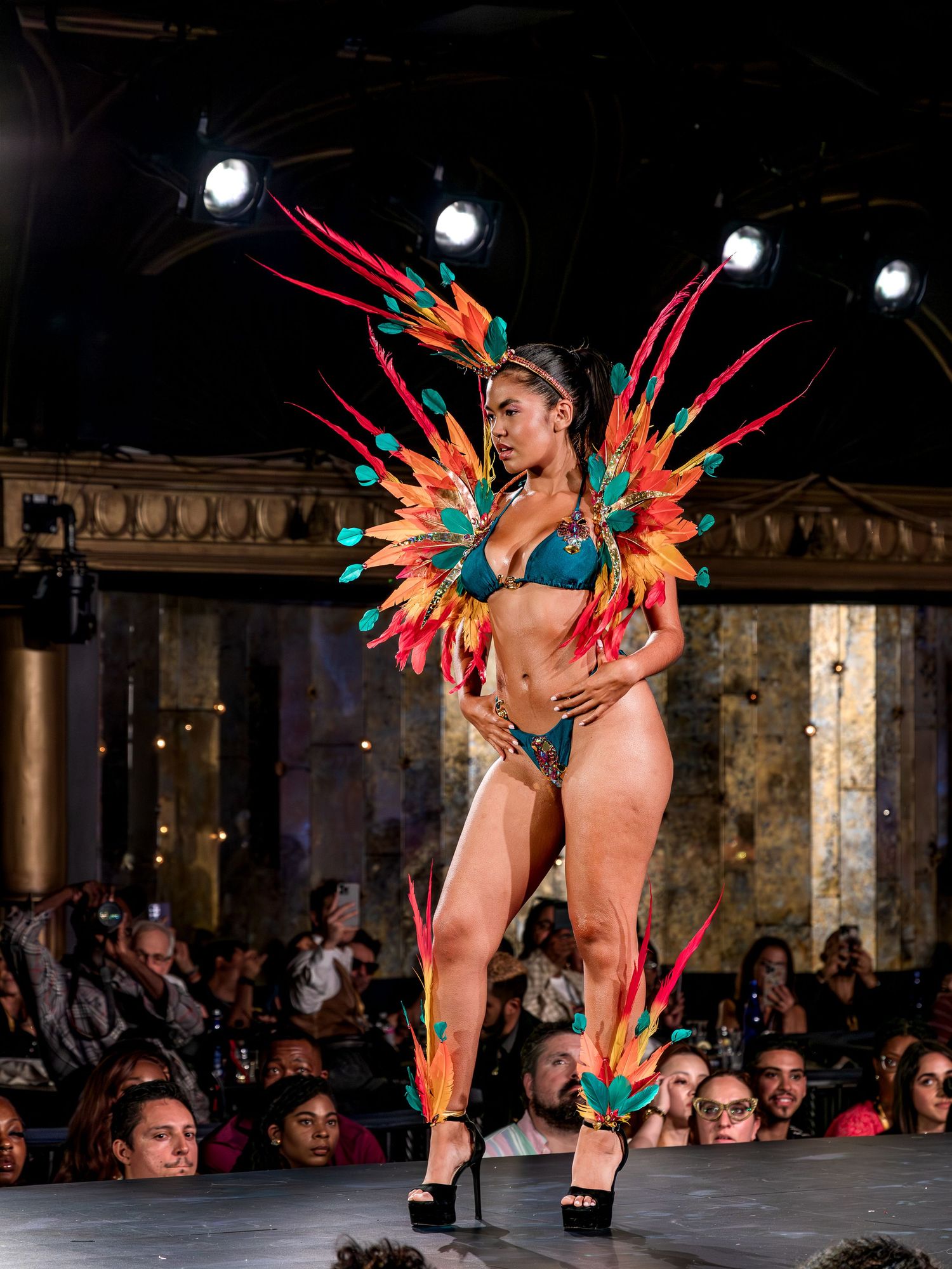 Model on runway in teal bikini with vibrant multicolor feather wing costume on fashion stage.