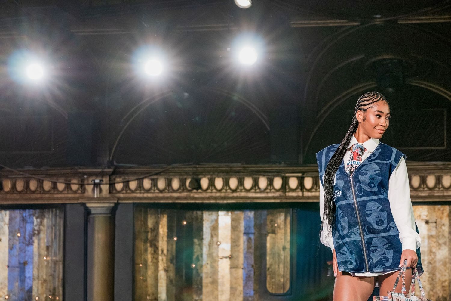 Female model walks runway in denim vest over white shirt with braided hairstyle and statement necklace.
