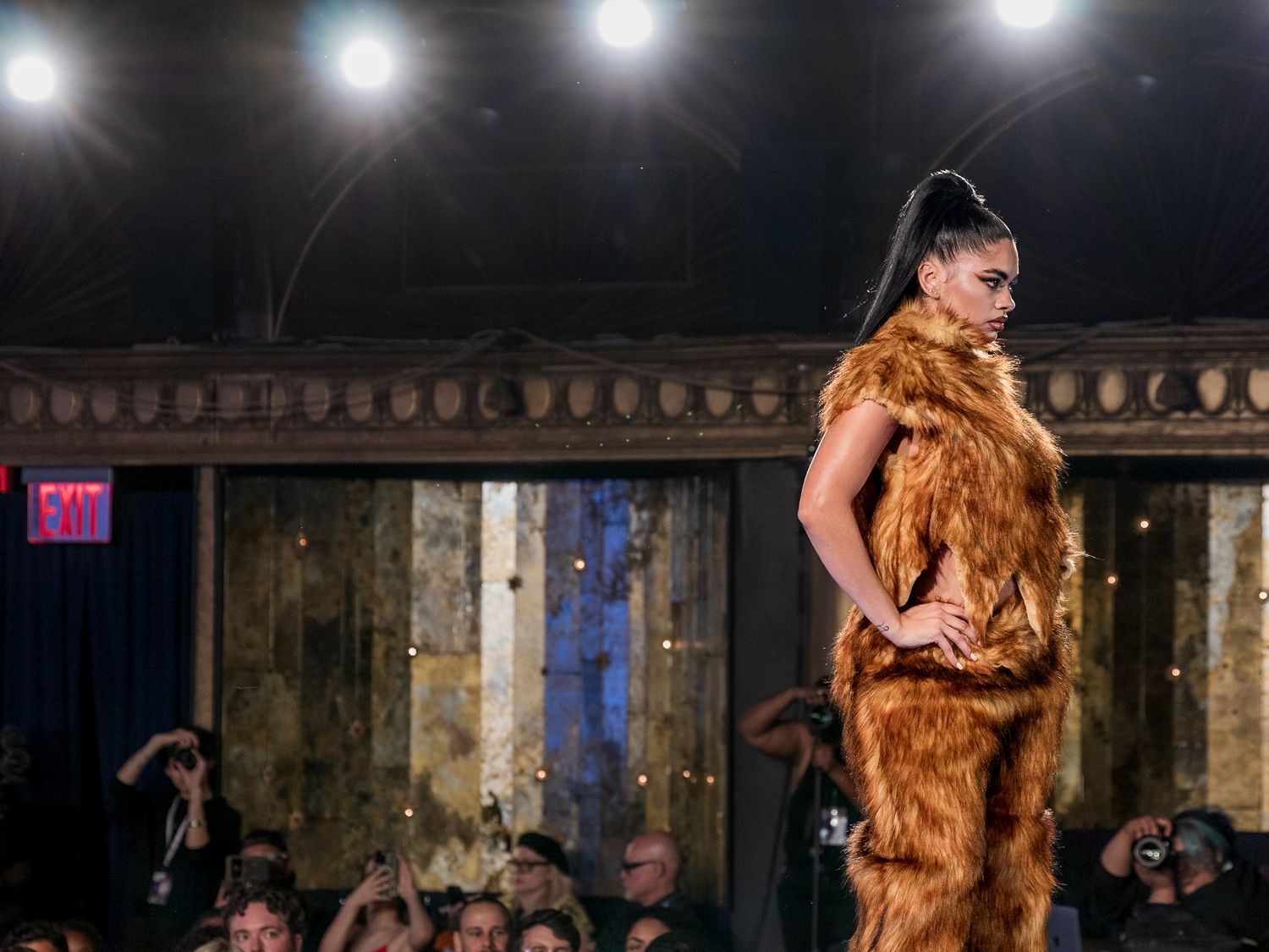 Model showcases sleek golden faux fur jumpsuit while walking runway at indoor fashion show.
